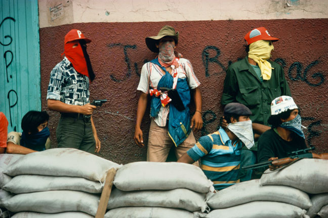 Colourful scene of protesters standing behind sandbag barricade, wearing protective headgear, in front of graffiti-covered wall.