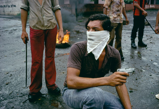 Masked protester with firearm in hand, standing on a street with other protesters in the background.
