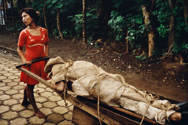 Woman in red top pulling sled with covered body through forest path.