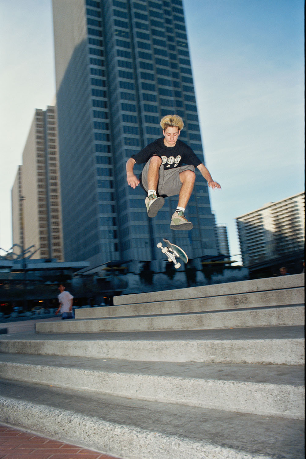 Skateboarder airborne above concrete steps with tall blue glass buildings and clear sky in background.