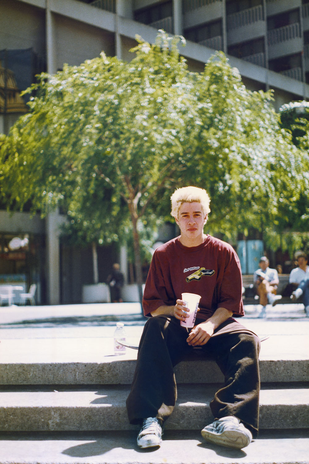 Young man with blonde hair sitting on concrete ledge holding drink, wearing burgundy top and dark trousers, large tree behind.