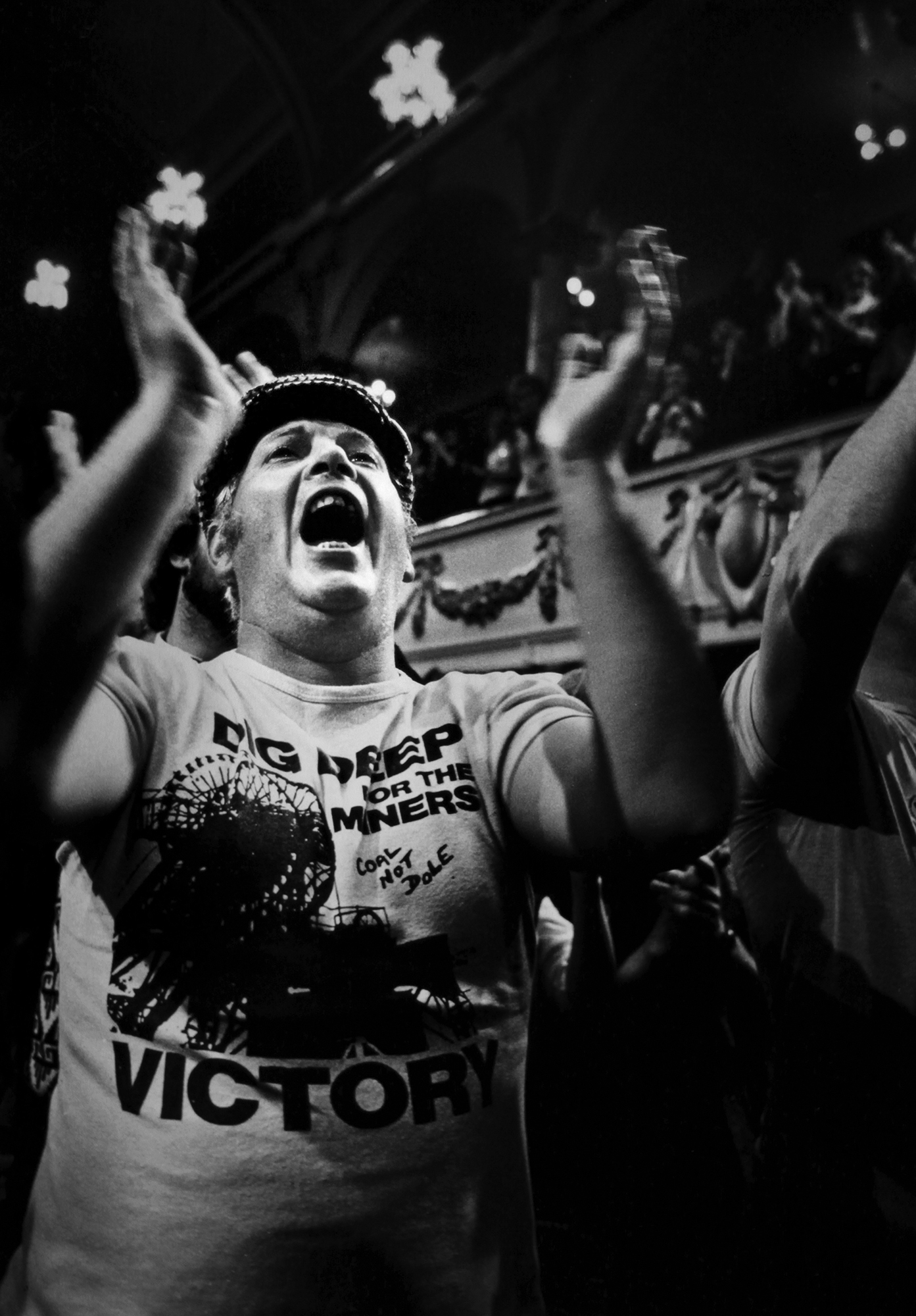 Black and white image of woman with raised arms celebrating, wearing "Victory" t-shirt and headband, surrounded by crowd indoors.