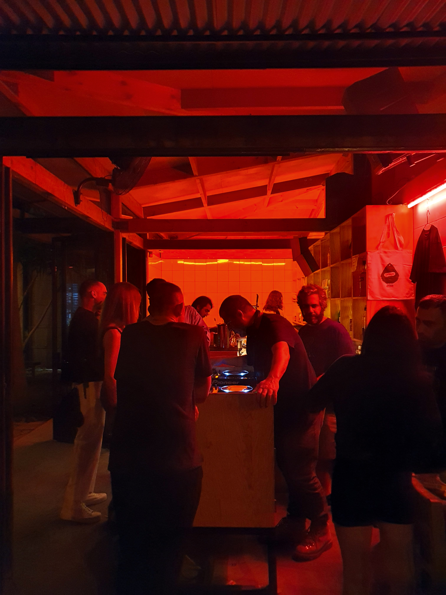 Crowded indoor space bathed in intense red lighting with exposed ceiling beams and silhouetted figures gathered around central area.