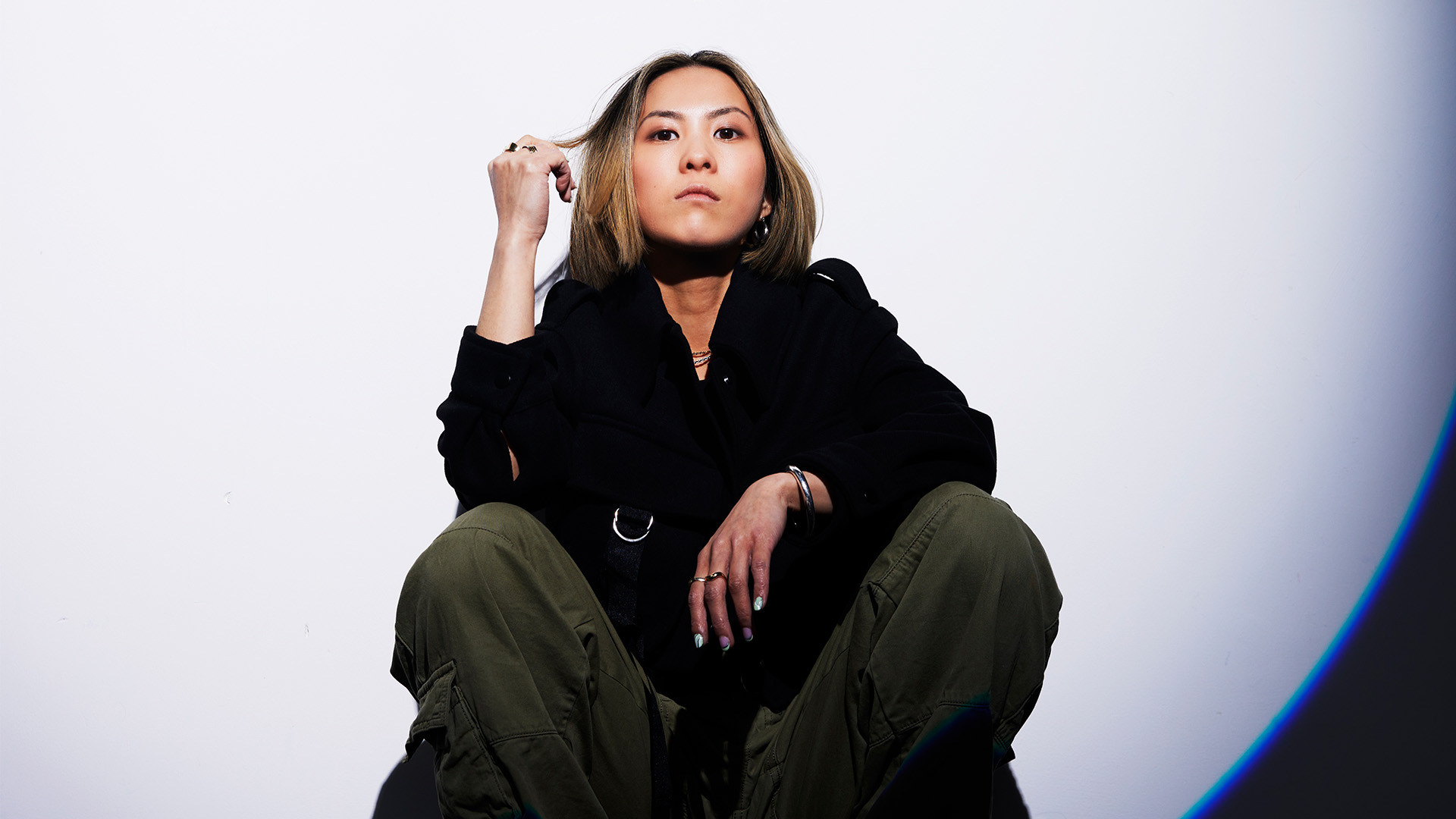 Woman in black hoodie and olive green trousers crouching against white backdrop, hand raised to head, looking upward.