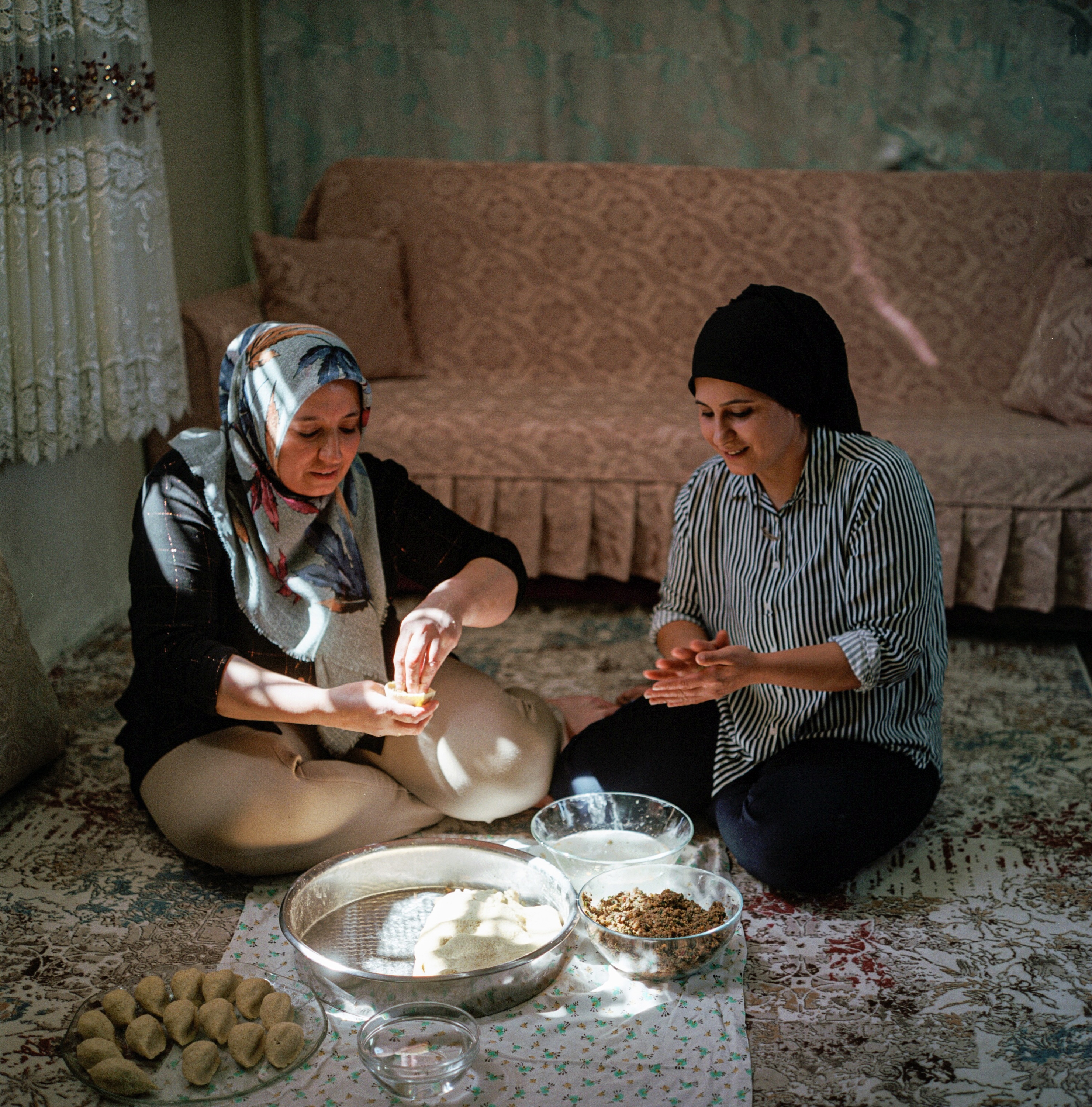 Two women in traditional clothing sitting on the floor, preparing food on a table with various dishes and ingredients.