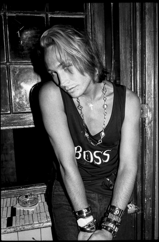 Woman with blonde hair wearing dark vest with "BOSS" text, chunky necklace and bracelets, leaning against wooden doorframe in black and white.