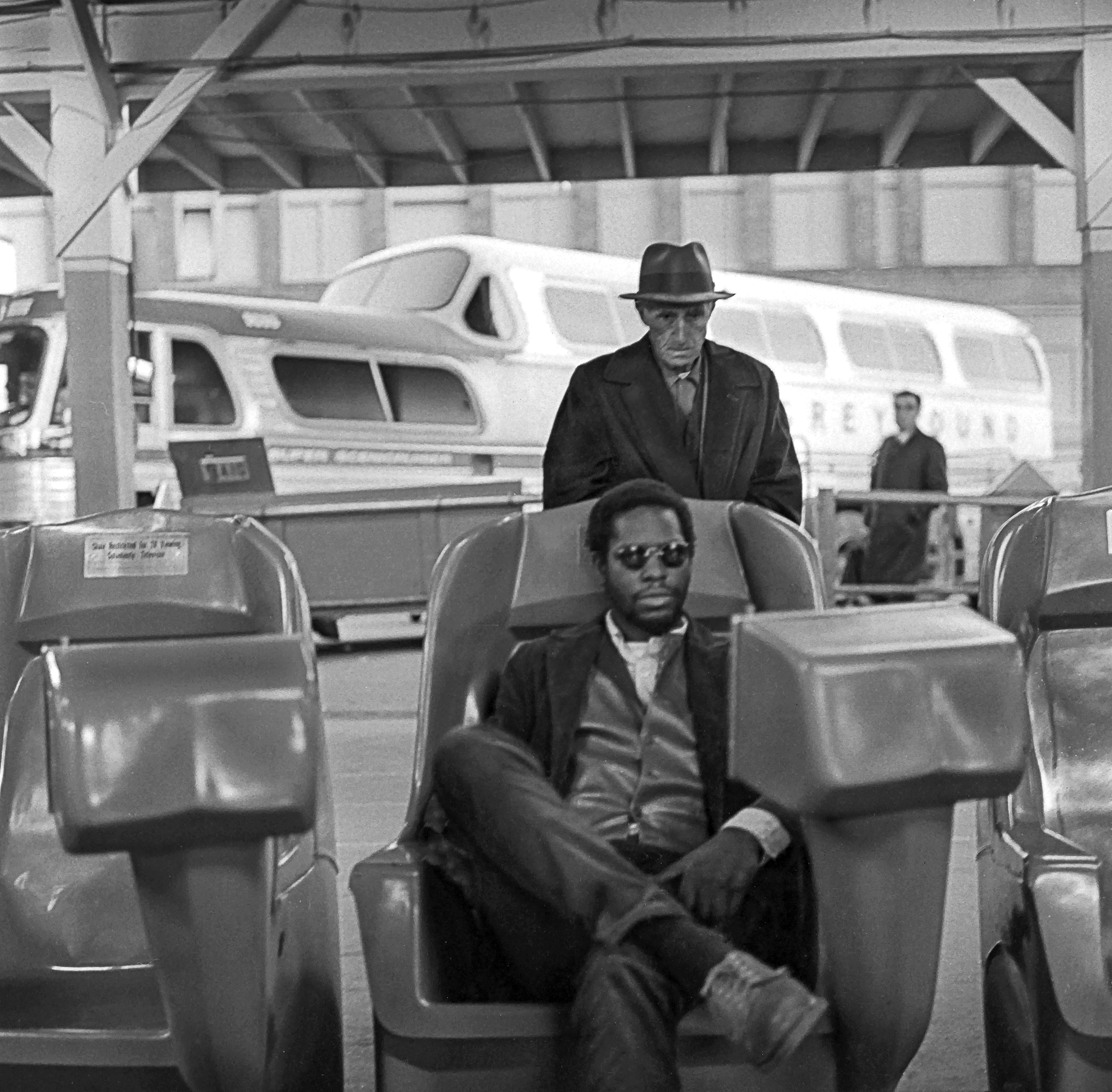 Two men seated on train platform benches, one standing and one sitting, in black and white photograph.