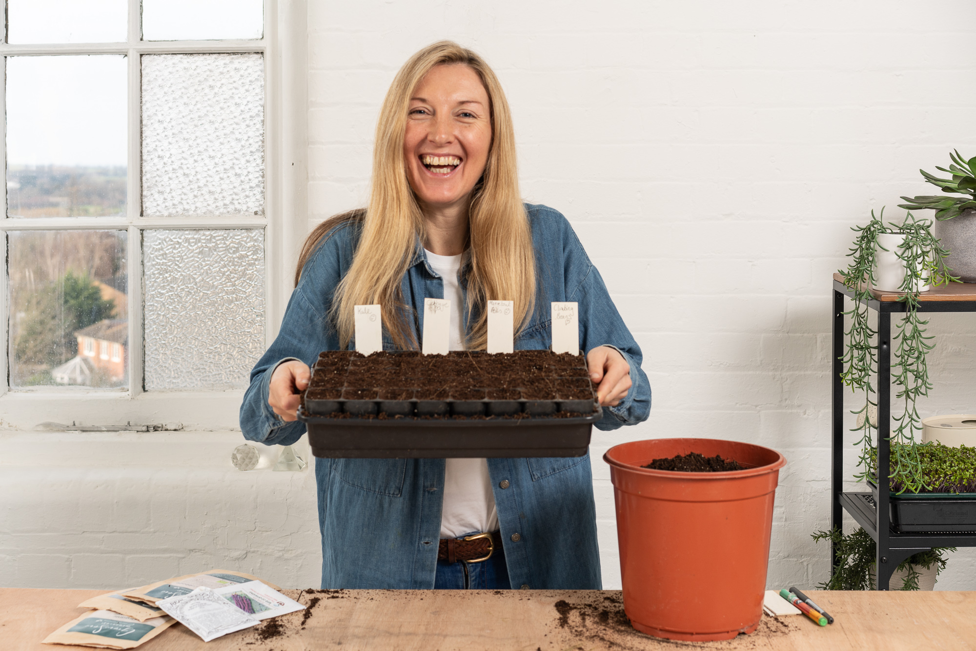 Blonde woman in blue denim jacket laughing whilst holding seed tray with white plant pots. Orange terracotta pot and gardening supplies on wooden table.