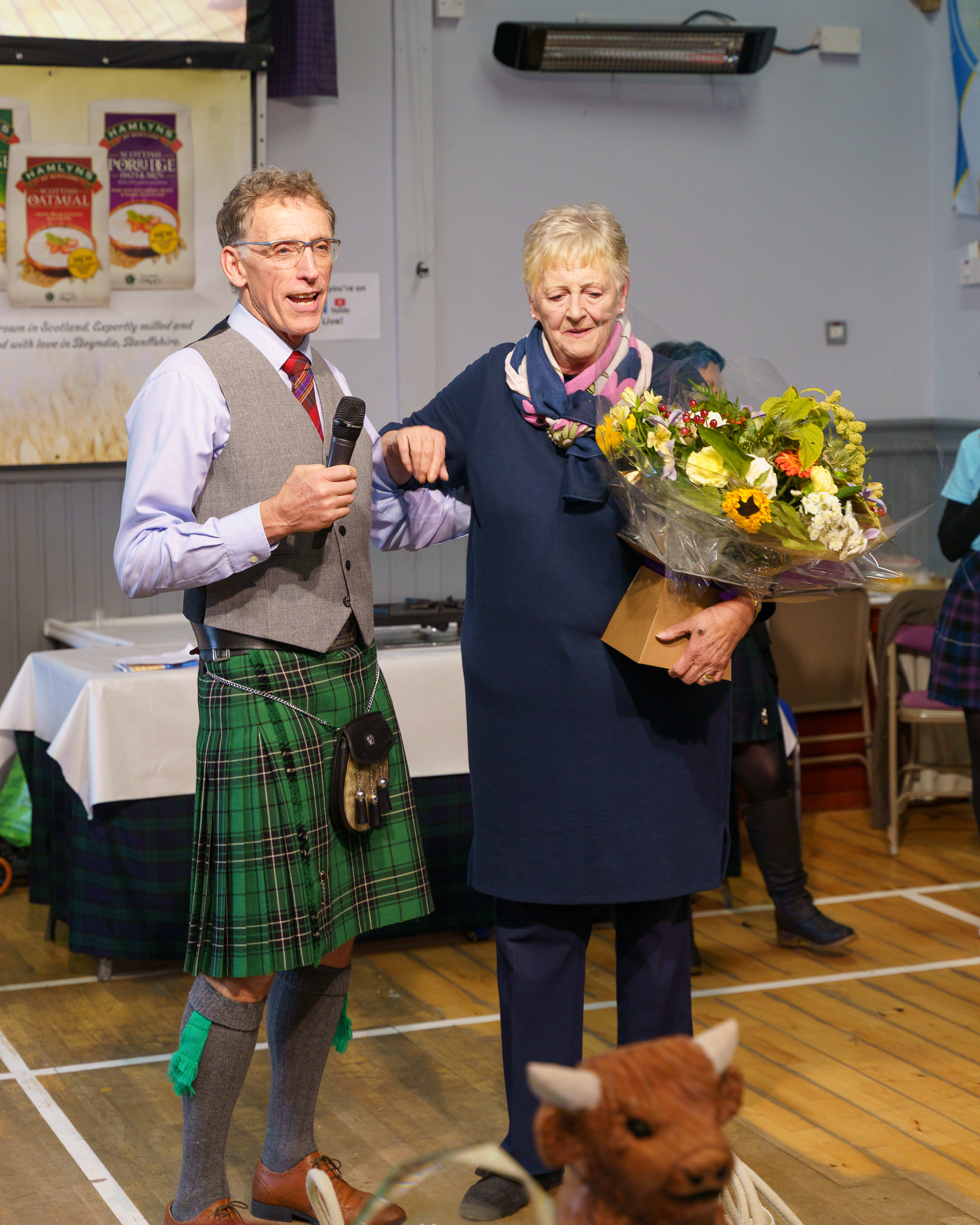Man in green tartan kilt and waistcoat holds microphone beside woman in navy coat carrying large floral bouquet in community hall.