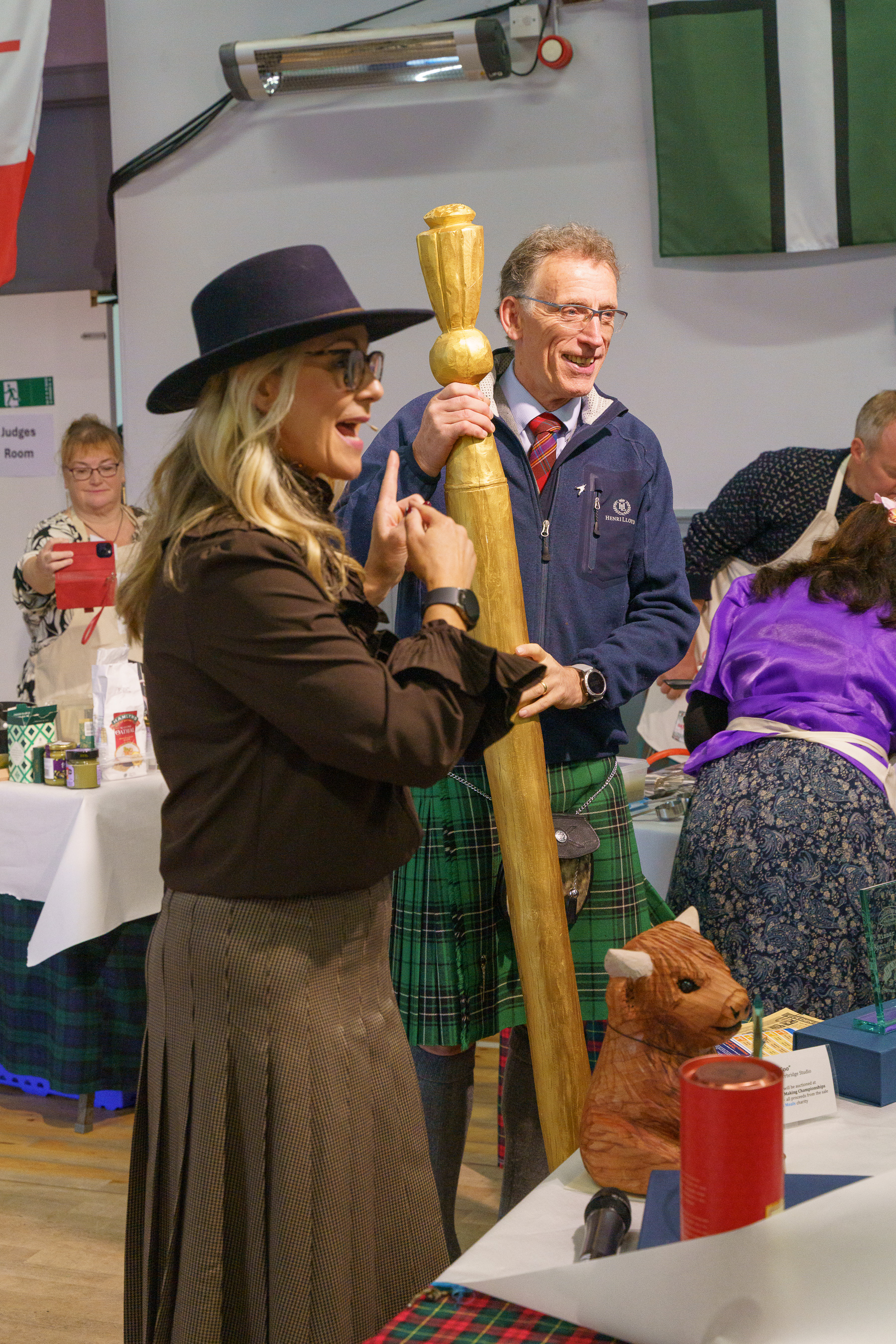 Woman in black hat and brown coat holds wooden staff beside man in blue jacket and green tartan kilt at indoor event with seated audience.