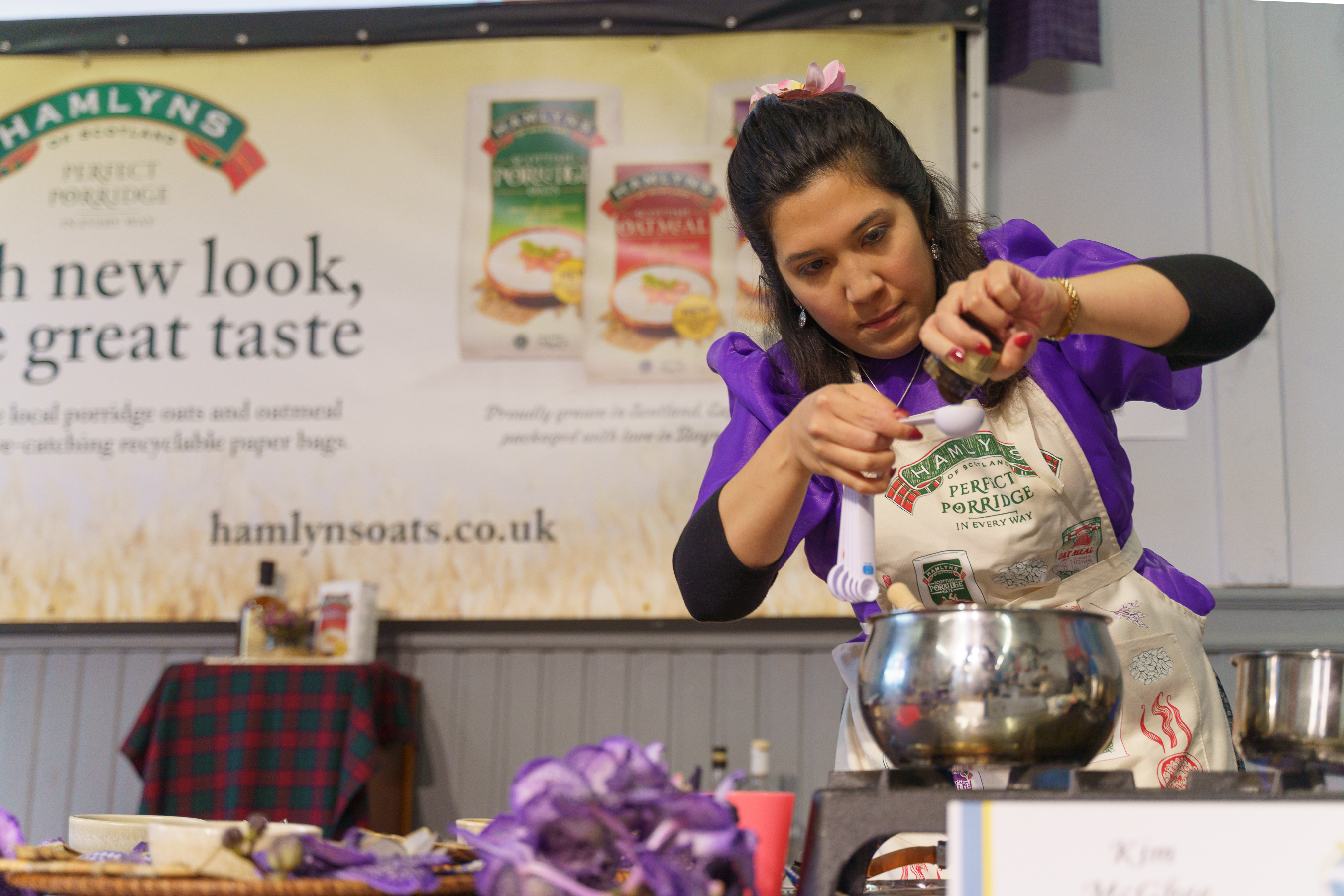 Woman in purple top and apron pouring ingredients into metal bowl at cooking demonstration, Hamlyns banner visible behind