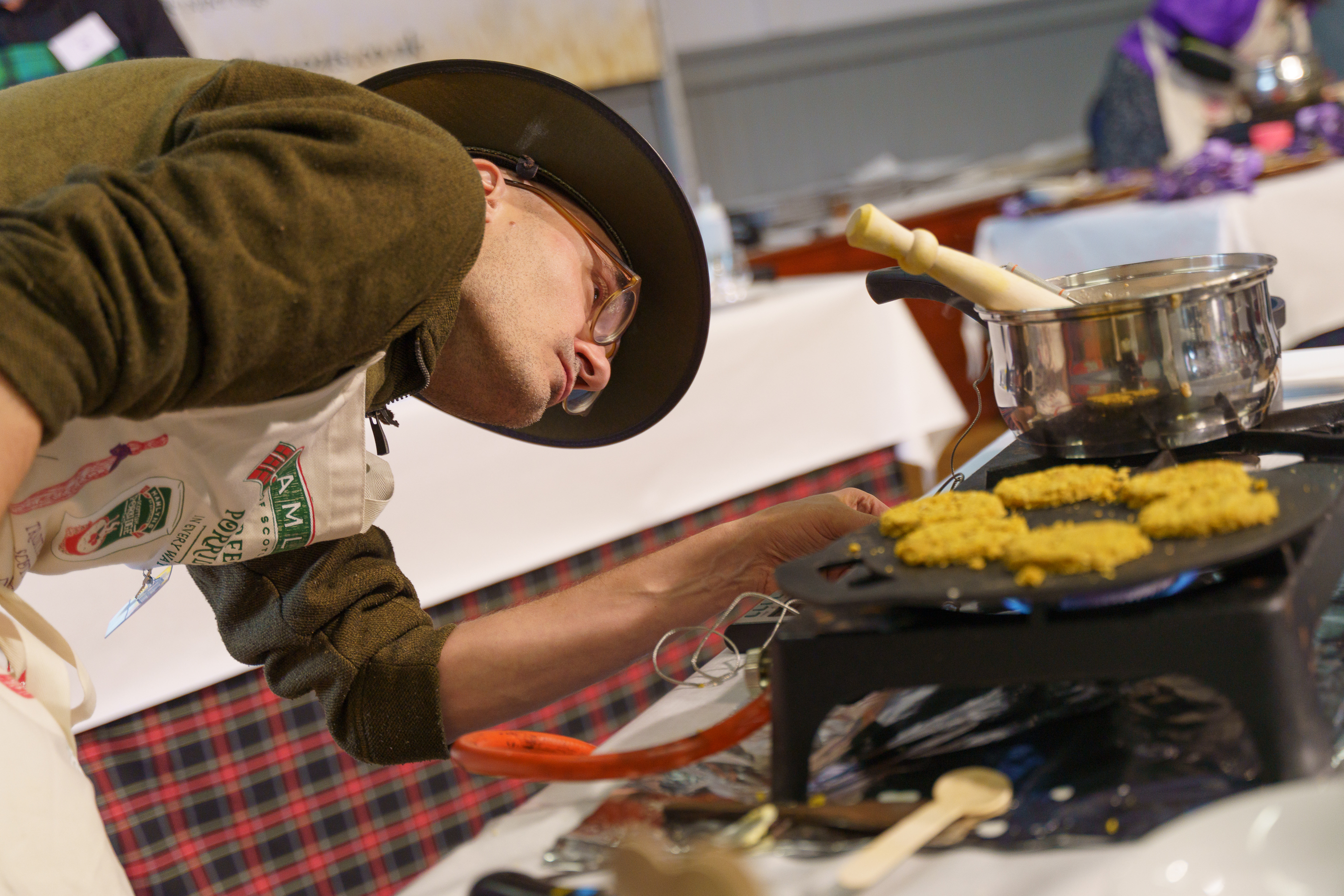 Person in green hoodie and black beanie cooking at stovetop with pan of yellow food and metal pot with wooden handle.