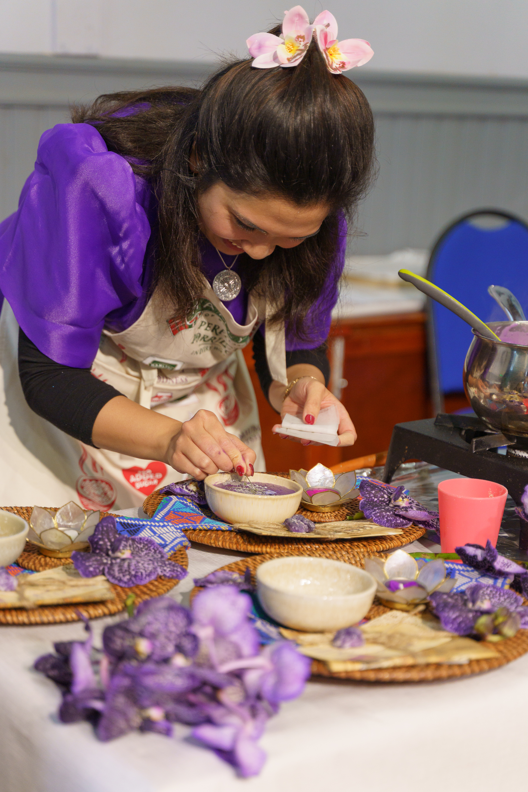 Woman in purple top with pink flower hair clip decorating cupcakes at table with purple flowers and baking supplies.