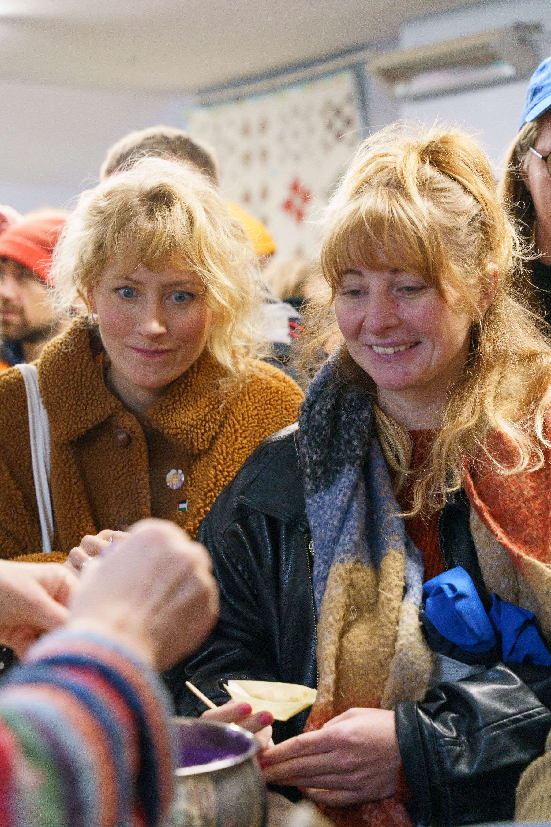 Two blonde women with fringes signing autographs, one in brown jacket, one in dark coat with scarf, crowded indoor setting.