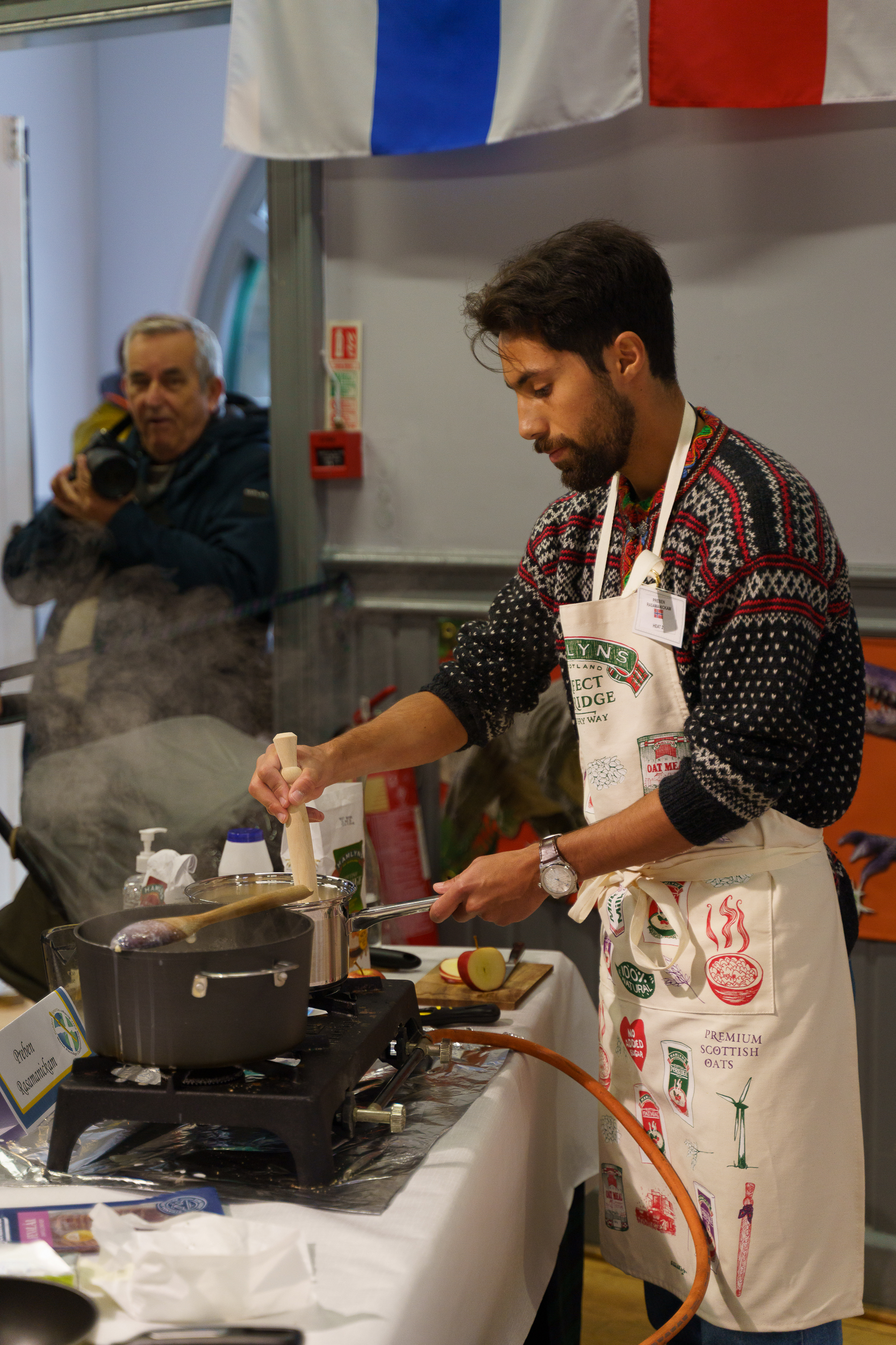 Man in patterned jumper and apron cooking at hob in kitchen, older man on phone in background, bunting overhead