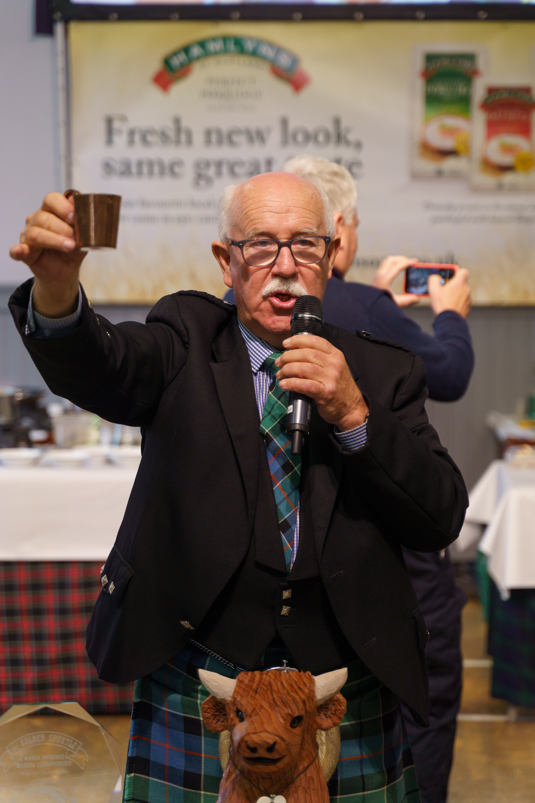 Elderly man in tartan kilt and dark jacket holds microphone and mug, speaking at event with branded backdrop behind him.