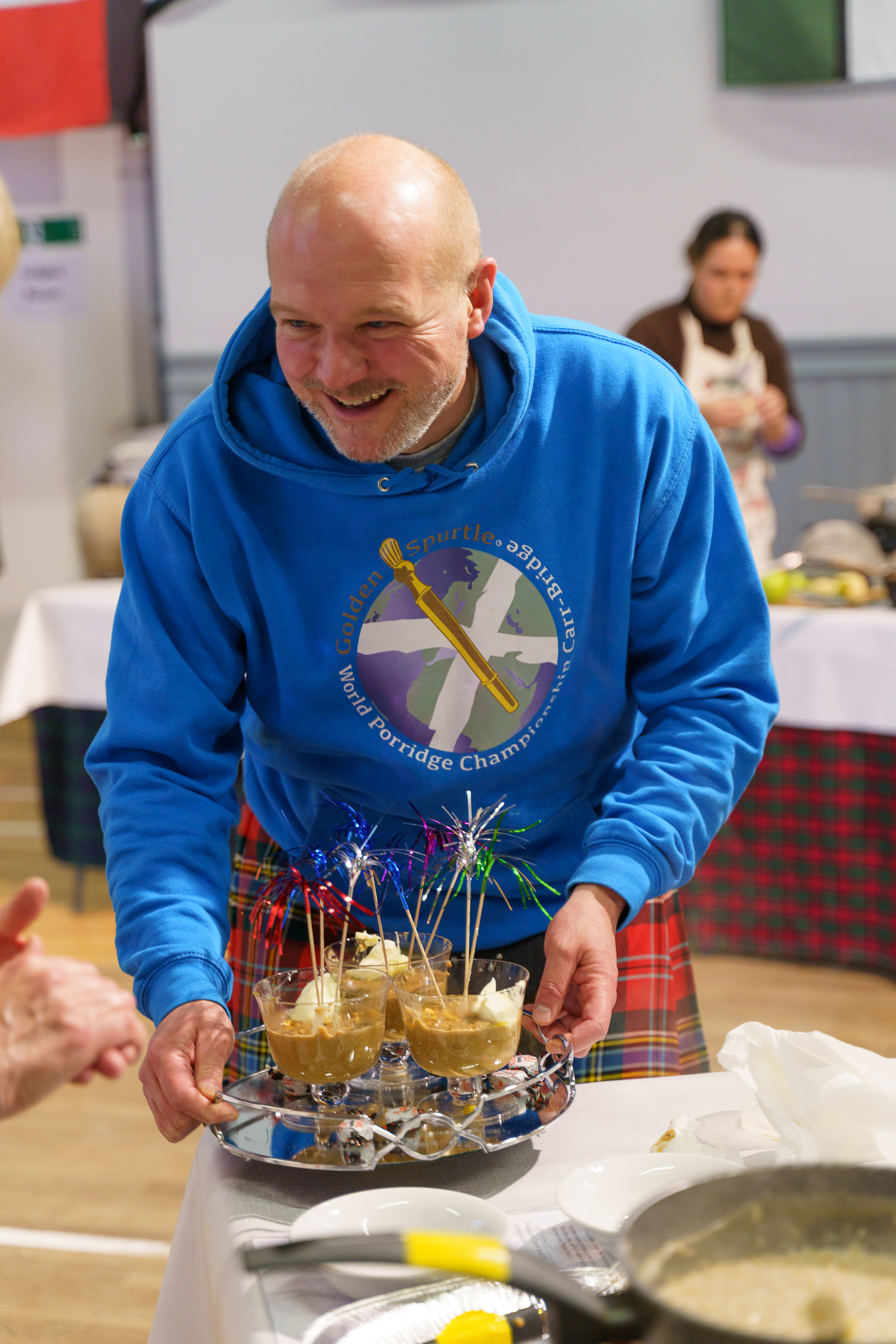 Bald man in blue hoodie holding tray with haggis portions topped with sparklers, smiling in kitchen setting with tartan fabric visible.
