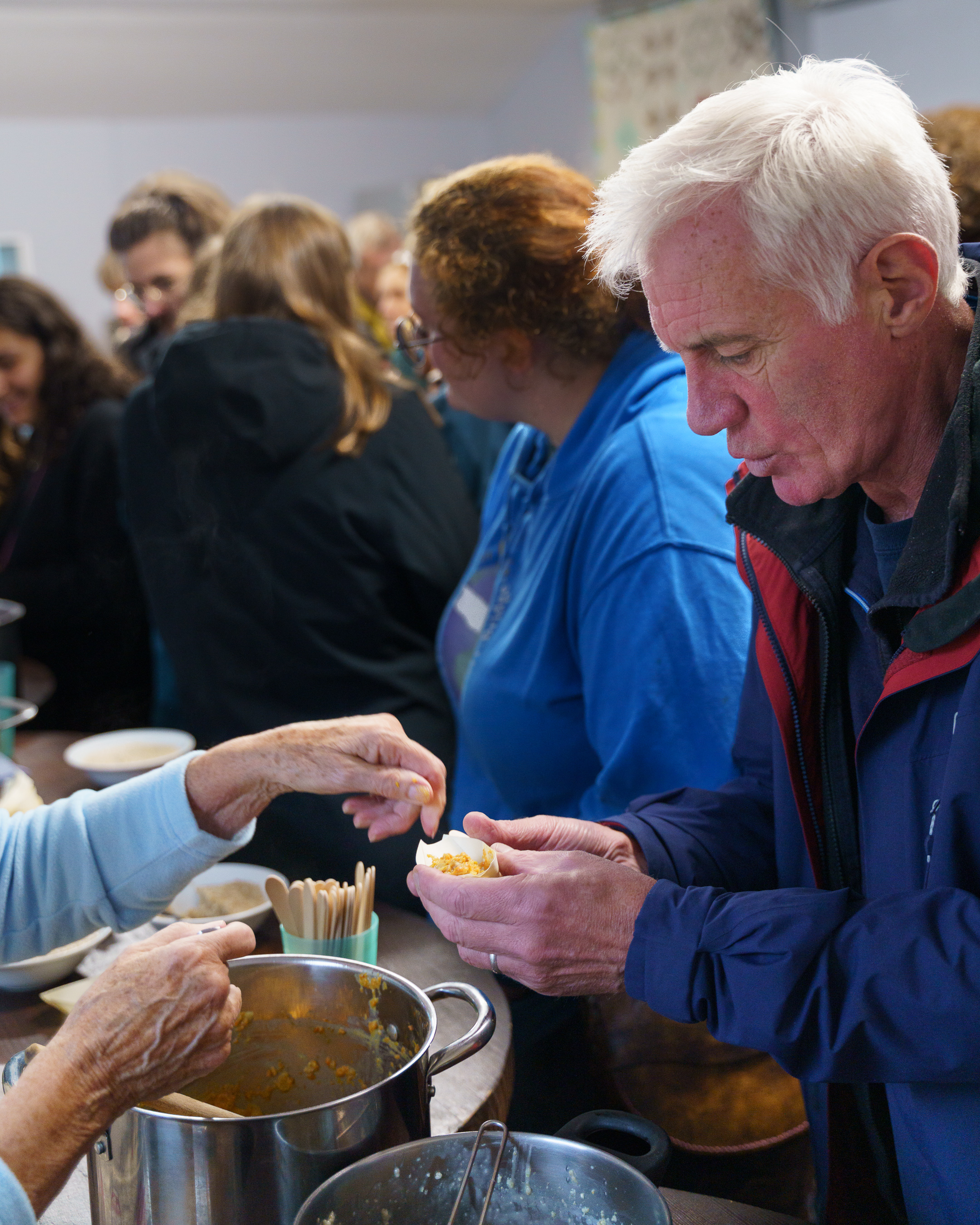 Elderly man in blue jacket being served food from large pot by person wearing light blue sleeve, with queue of people behind him.