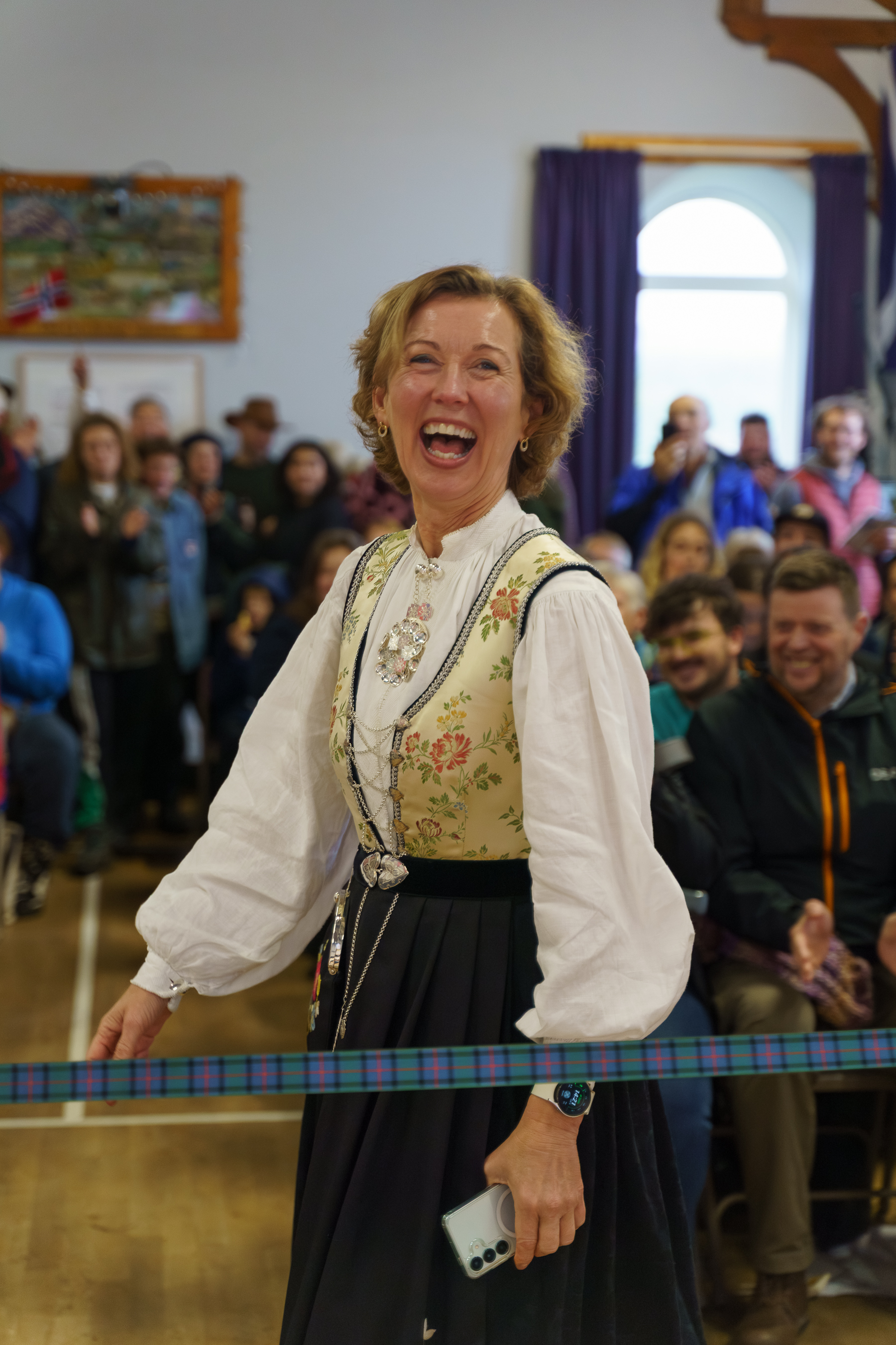 Woman in traditional dirndl with floral bodice and white blouse laughing at indoor event with seated audience in background.