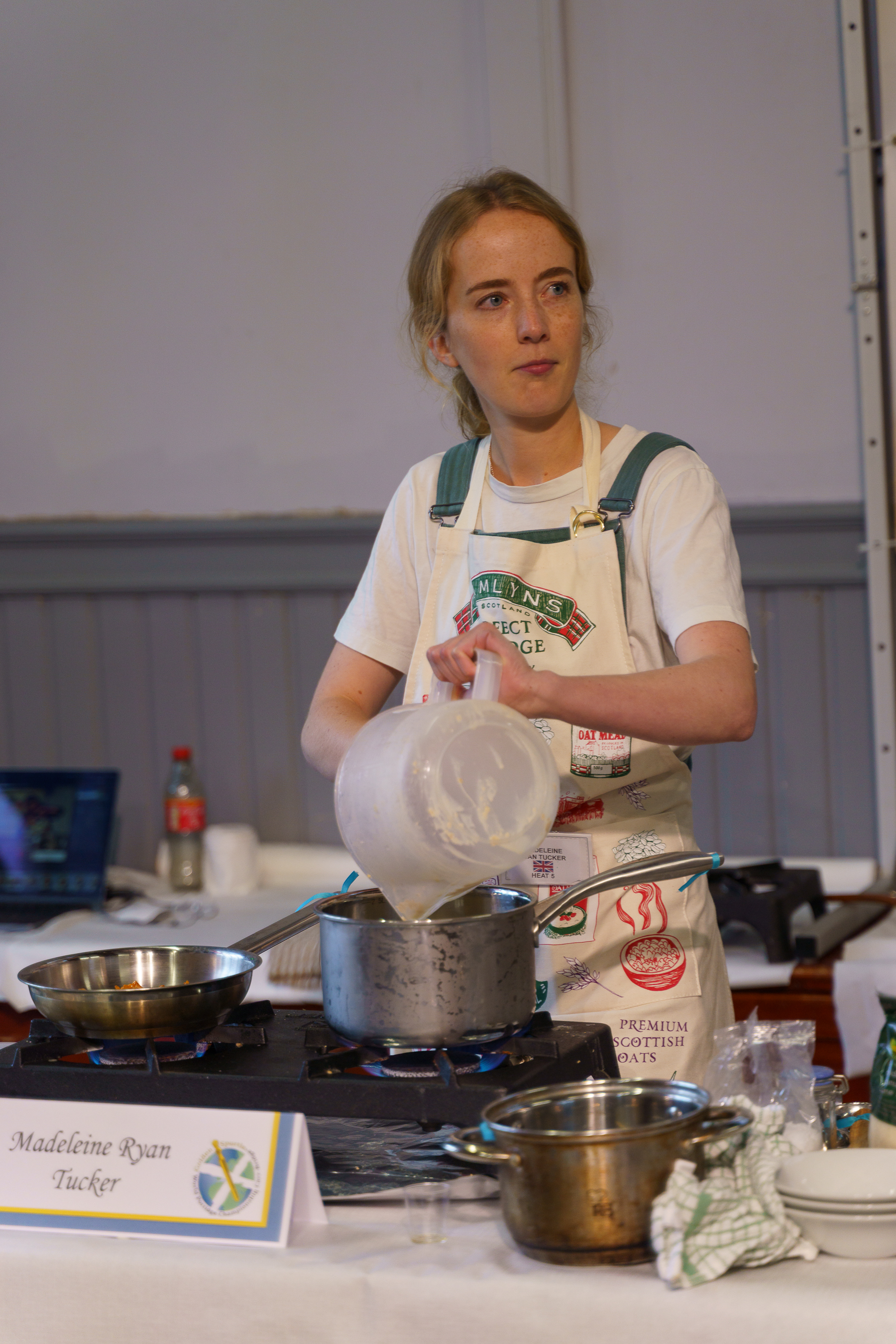 Woman in white apron cooking at demonstration kitchen with metal pans on hob, holding white bowl, name placard visible on counter.