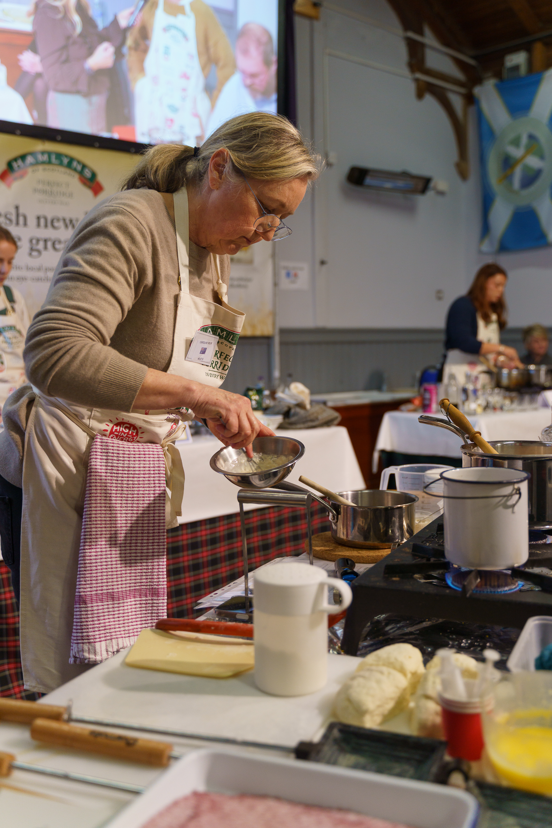 Woman in beige jumper and striped apron cooking at hob with saucepan, white mug and ingredients on counter at demonstration