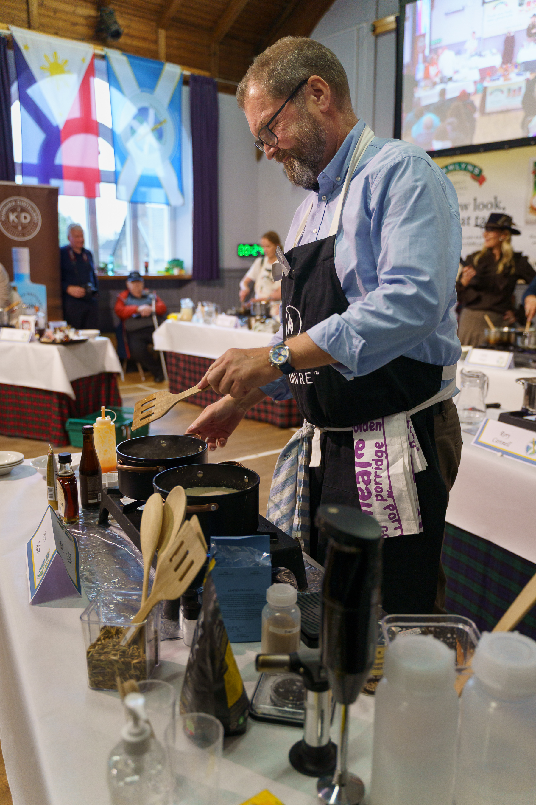 Chef in blue shirt and black apron cooking at stovetop during culinary demonstration, with cooking utensils and ingredients on counter.