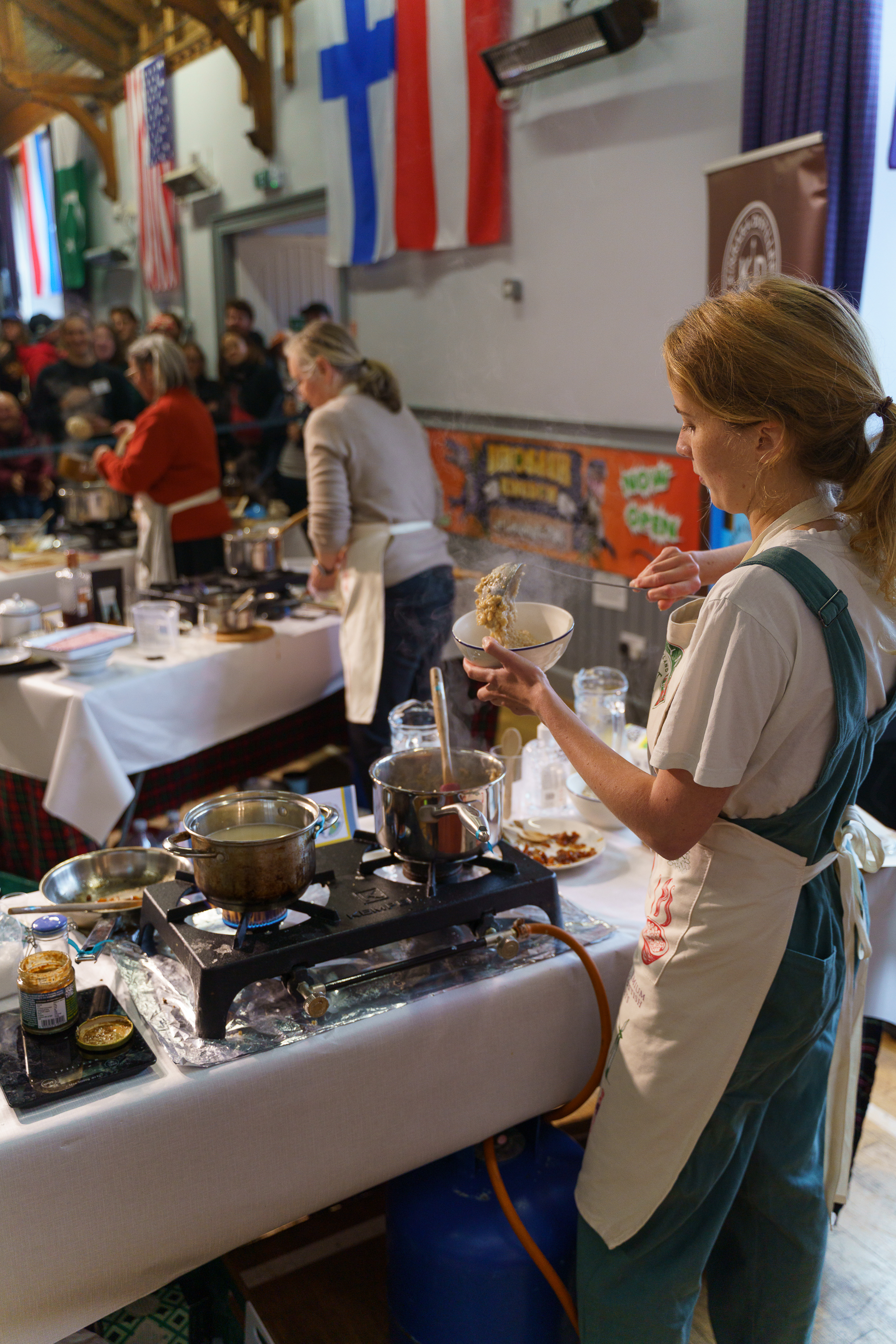Woman in green apron and white blouse cooking at portable hob on white table in hall with flags and seated audience watching.