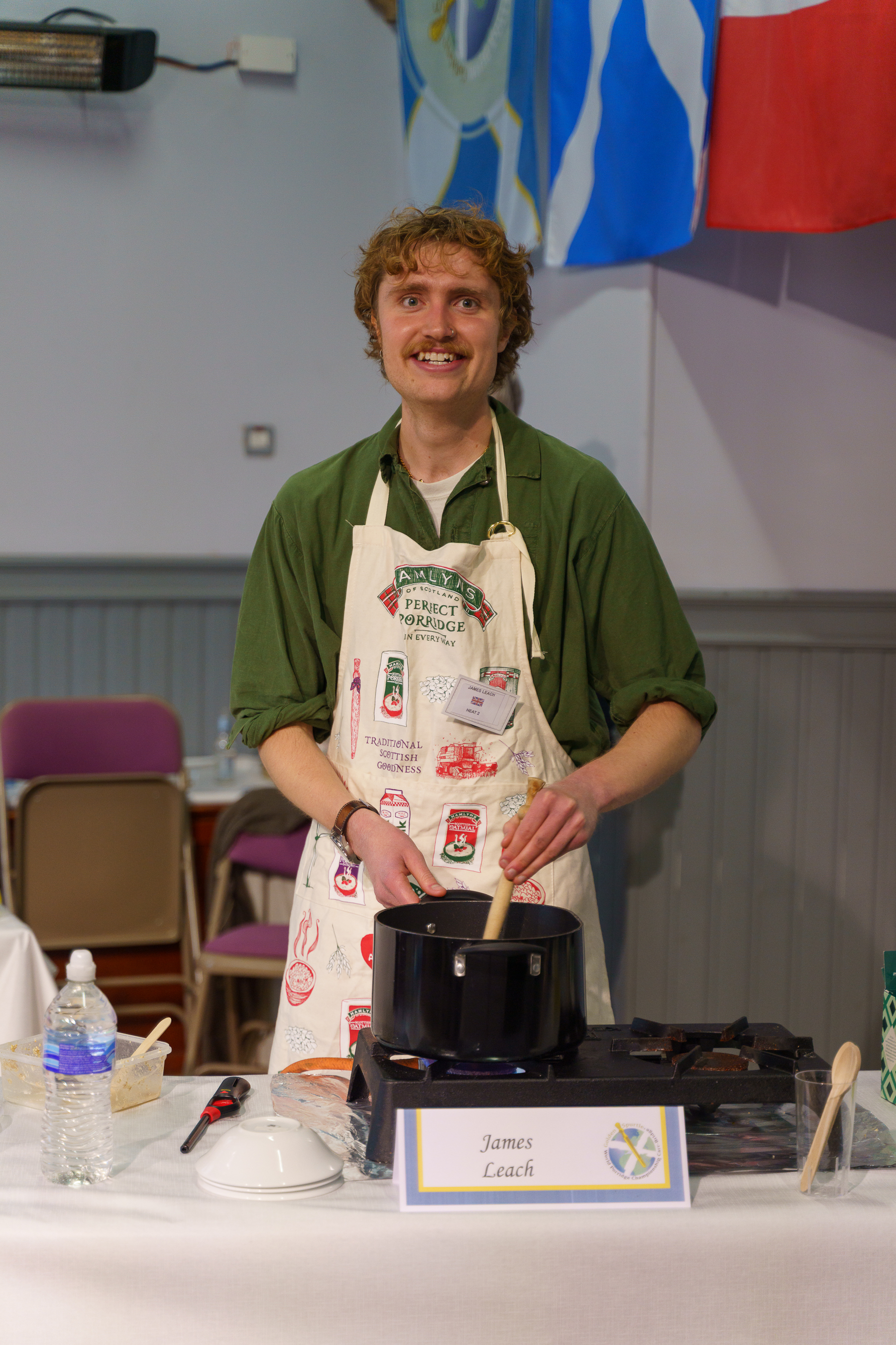 Man with curly hair and moustache wearing green shirt and cream apron stands behind cooking station with black pot and utensils.