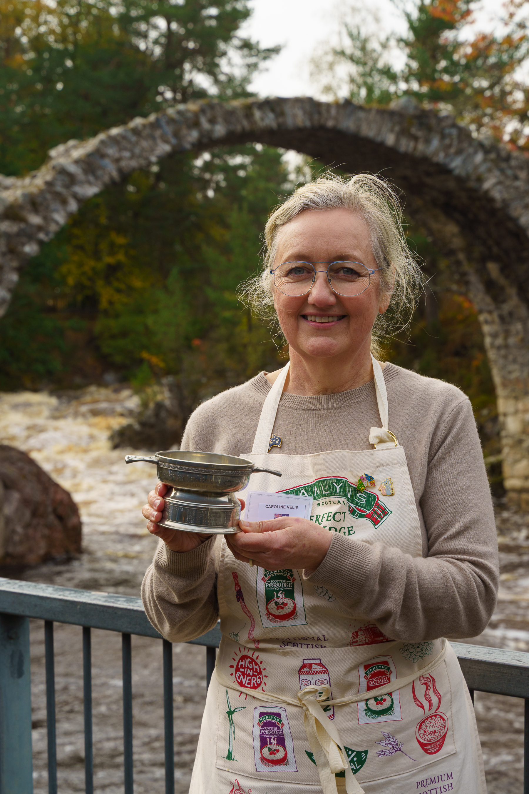 Woman with grey hair and glasses holding metal trophy, wearing beige apron with badges, standing by stone bridge and trees.