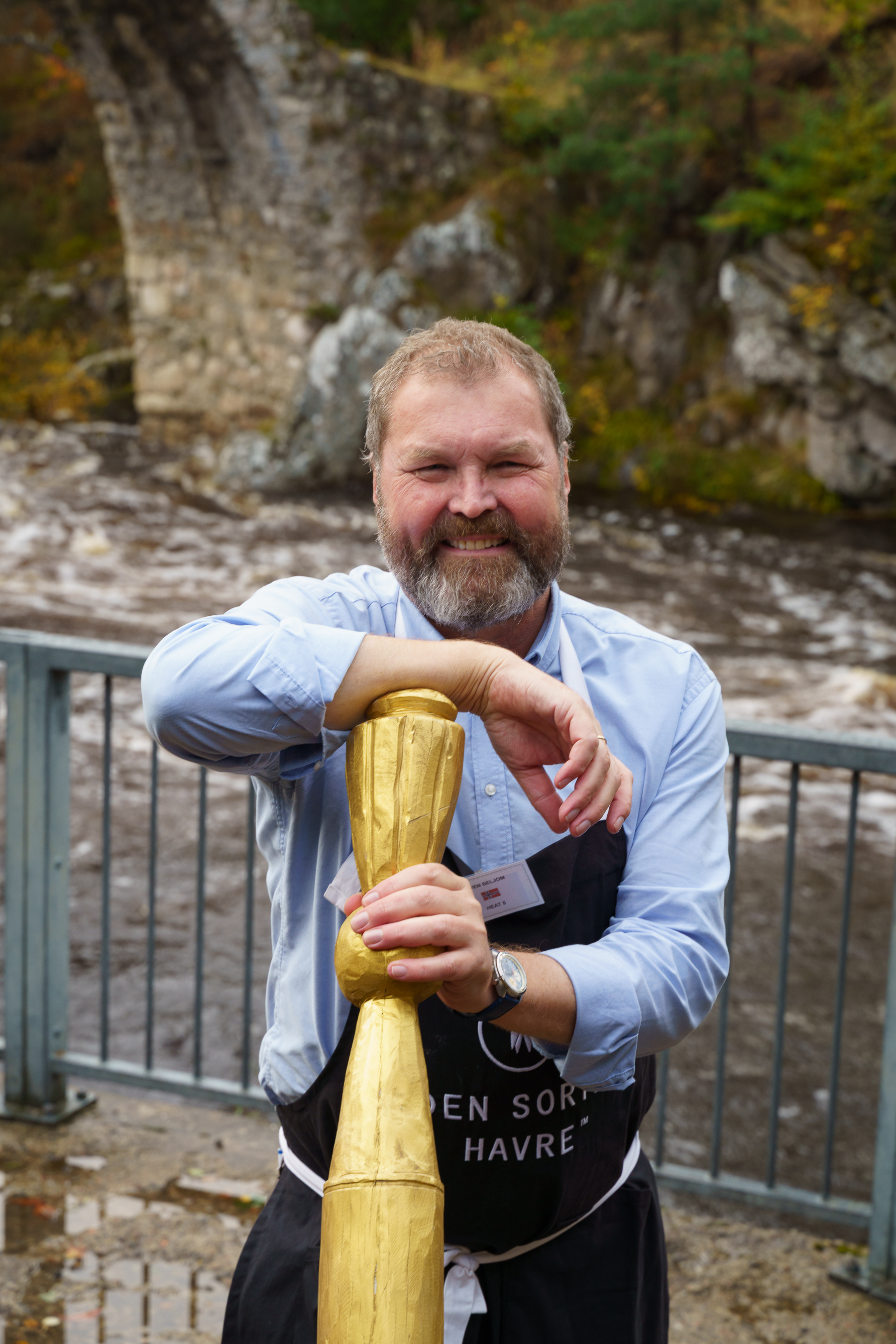 Smiling bearded man in light blue shirt and dark apron leaning on wooden post, stone wall and autumn foliage in background.