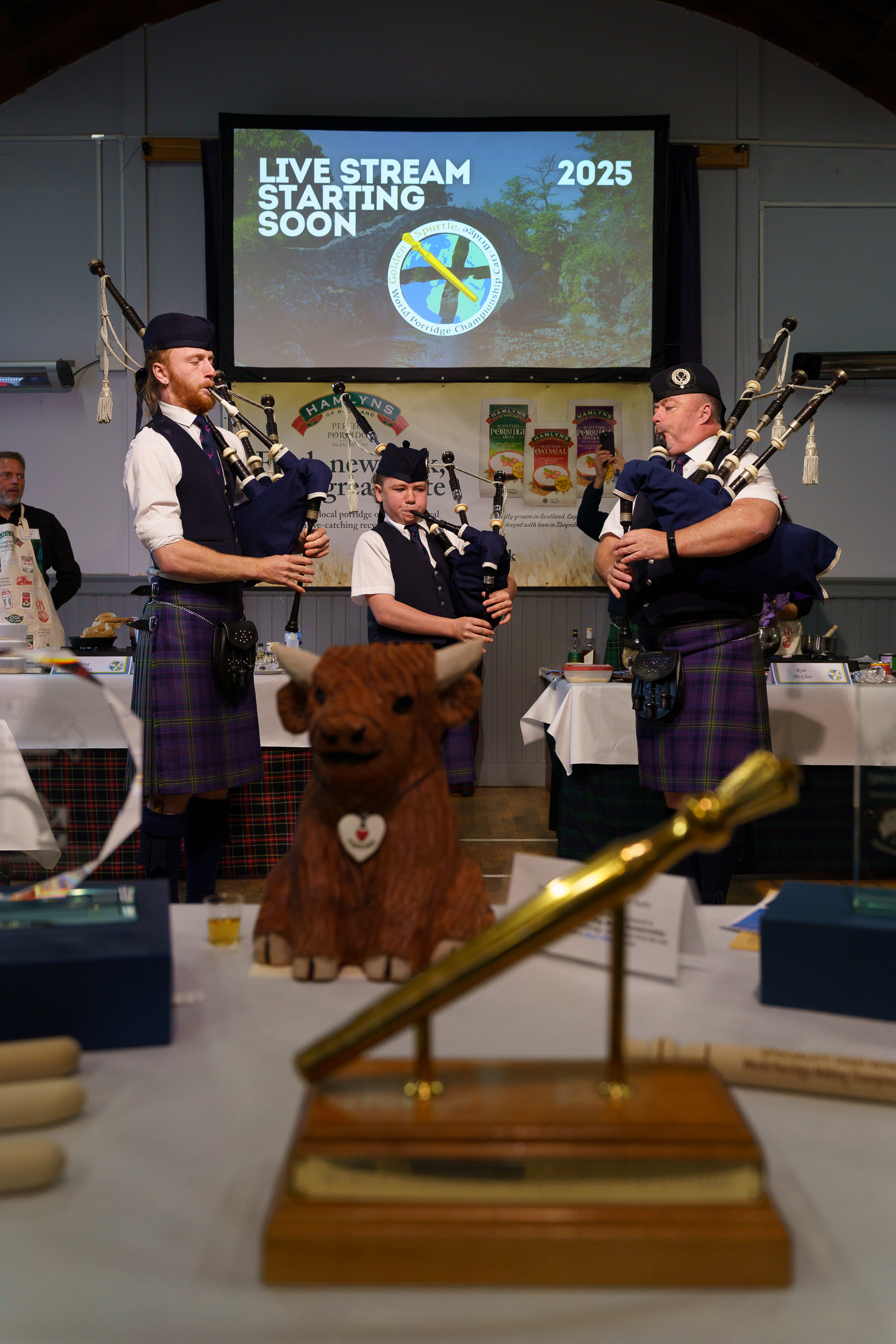 Three bagpipers in traditional Scottish dress performing indoors, with wooden Highland cow sculpture in foreground and projection screen behind.