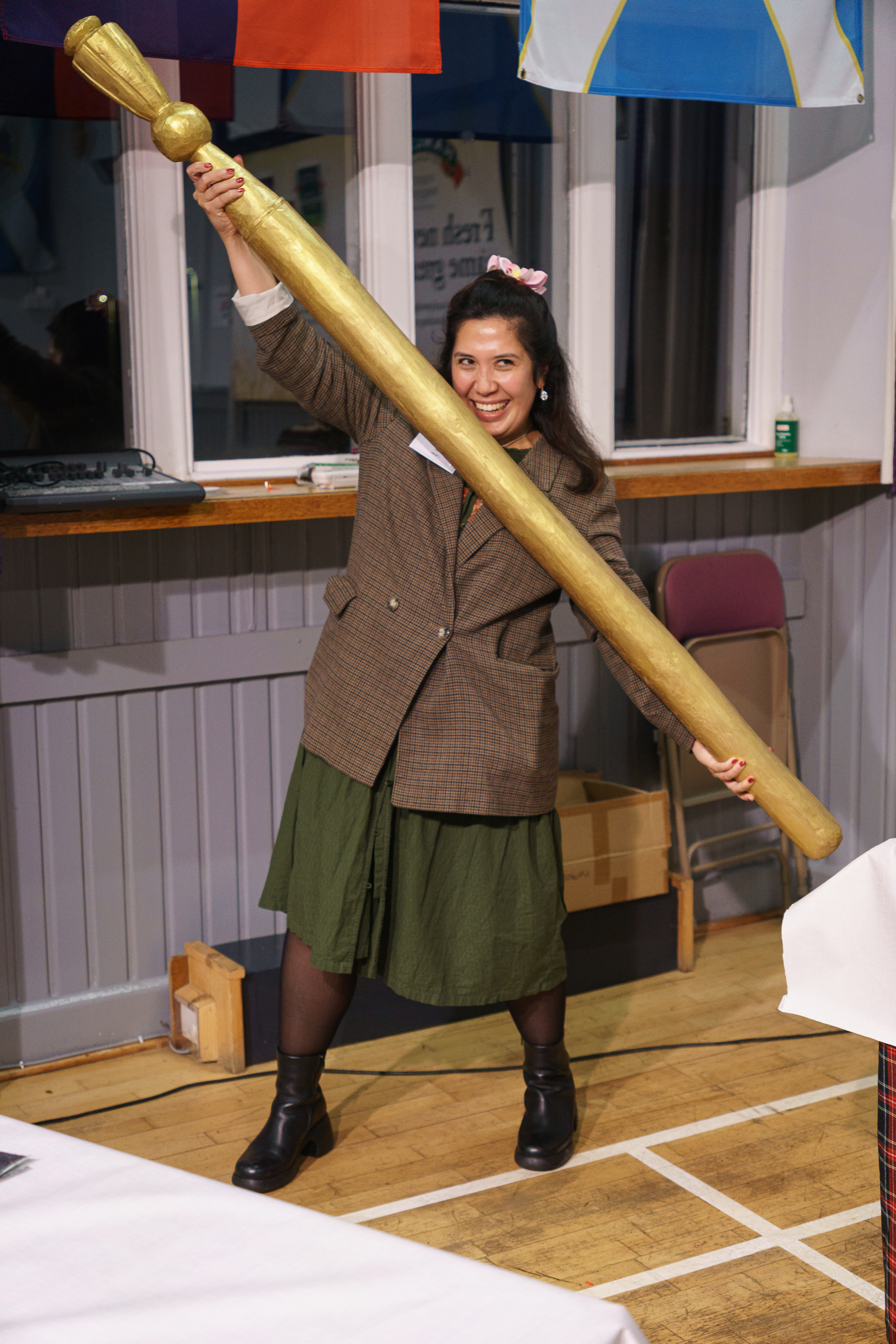 Woman in brown jacket and green skirt holds large golden rolling pin diagonally across body in kitchen setting with coloured bunting overhead.