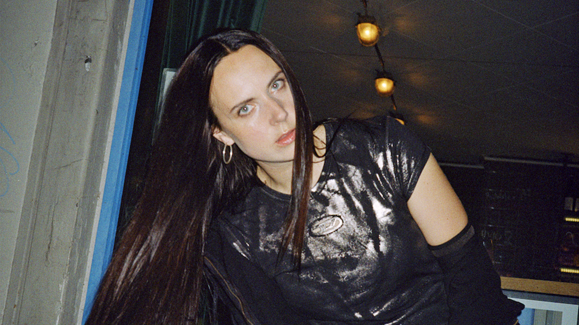 Woman with long dark hair wearing black leather jacket, sitting indoors with warm lighting and blue architectural elements visible.