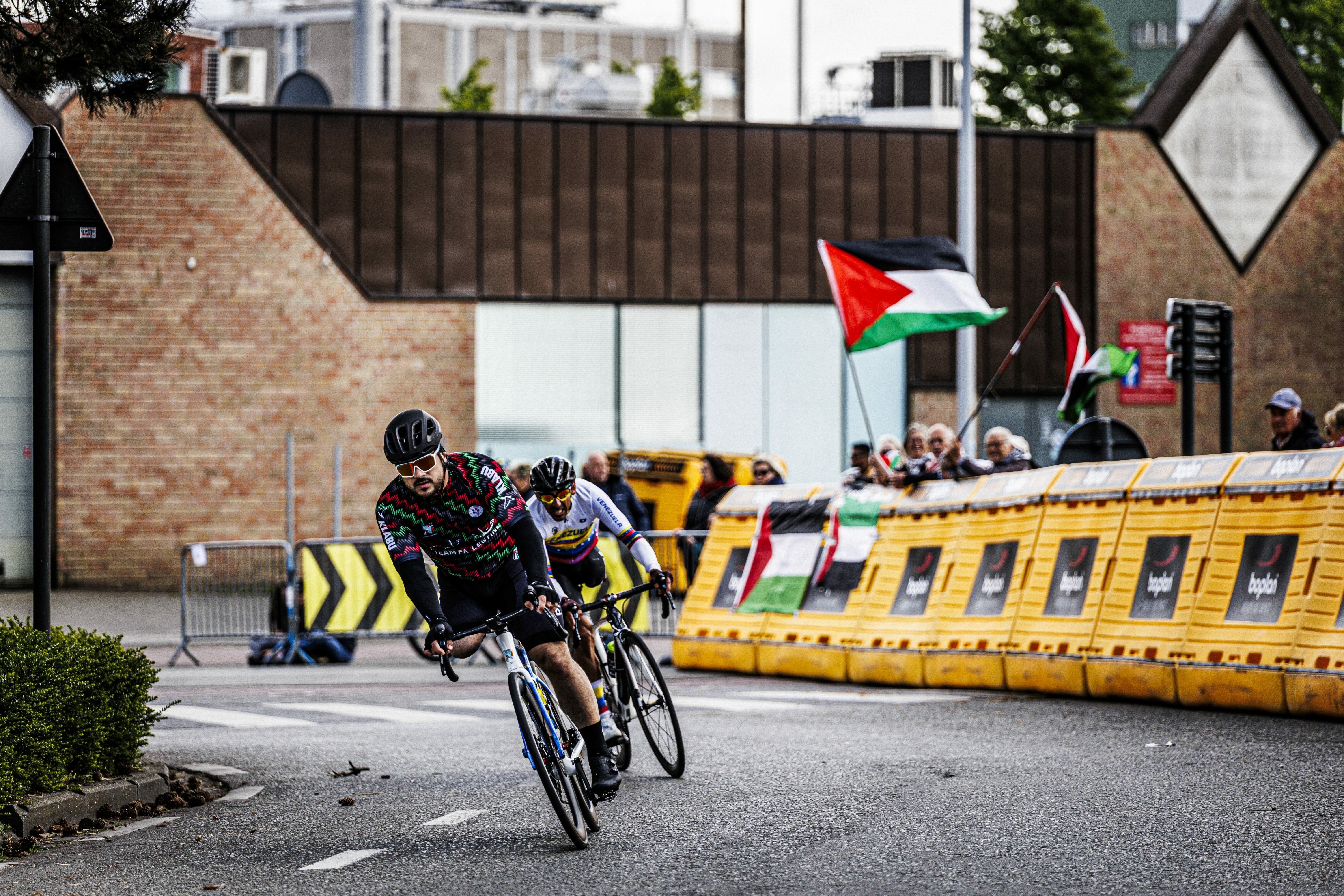 Cyclists racing past Palestinian flag, yellow barriers, and spectators.