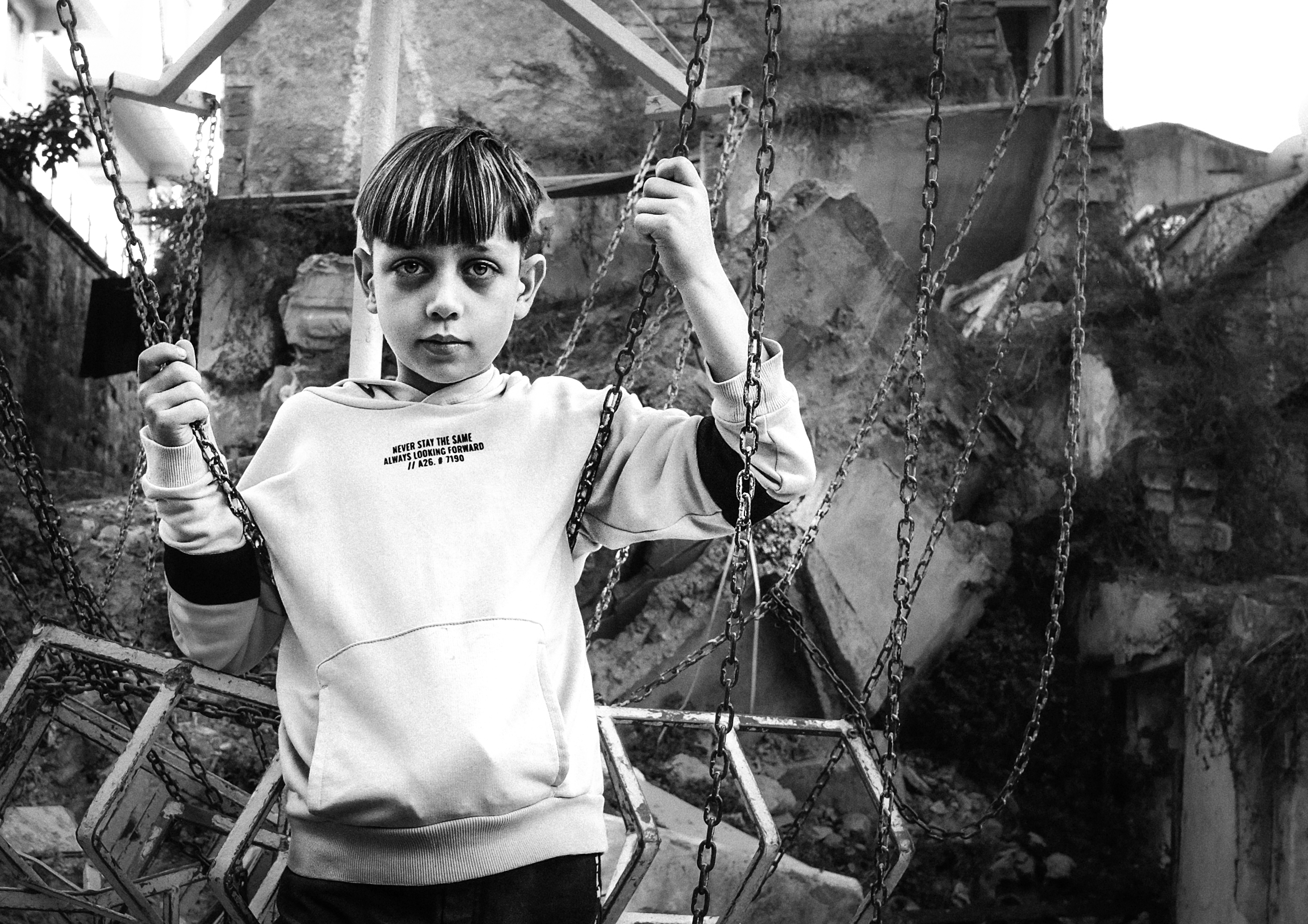A black and white photograph of a young boy standing on a swing set, with a rugged, dilapidated background.