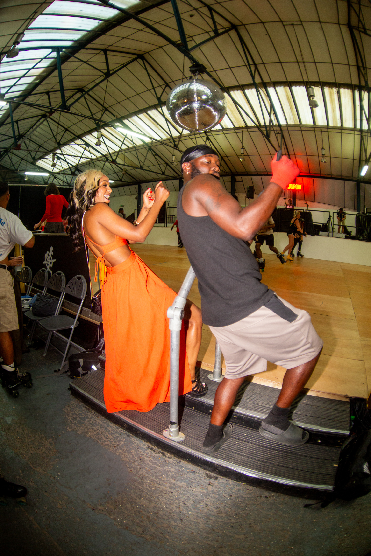 Two people dancing under arched glass roof structure. Woman in orange dress, man in grey vest and light shorts on raised platform.