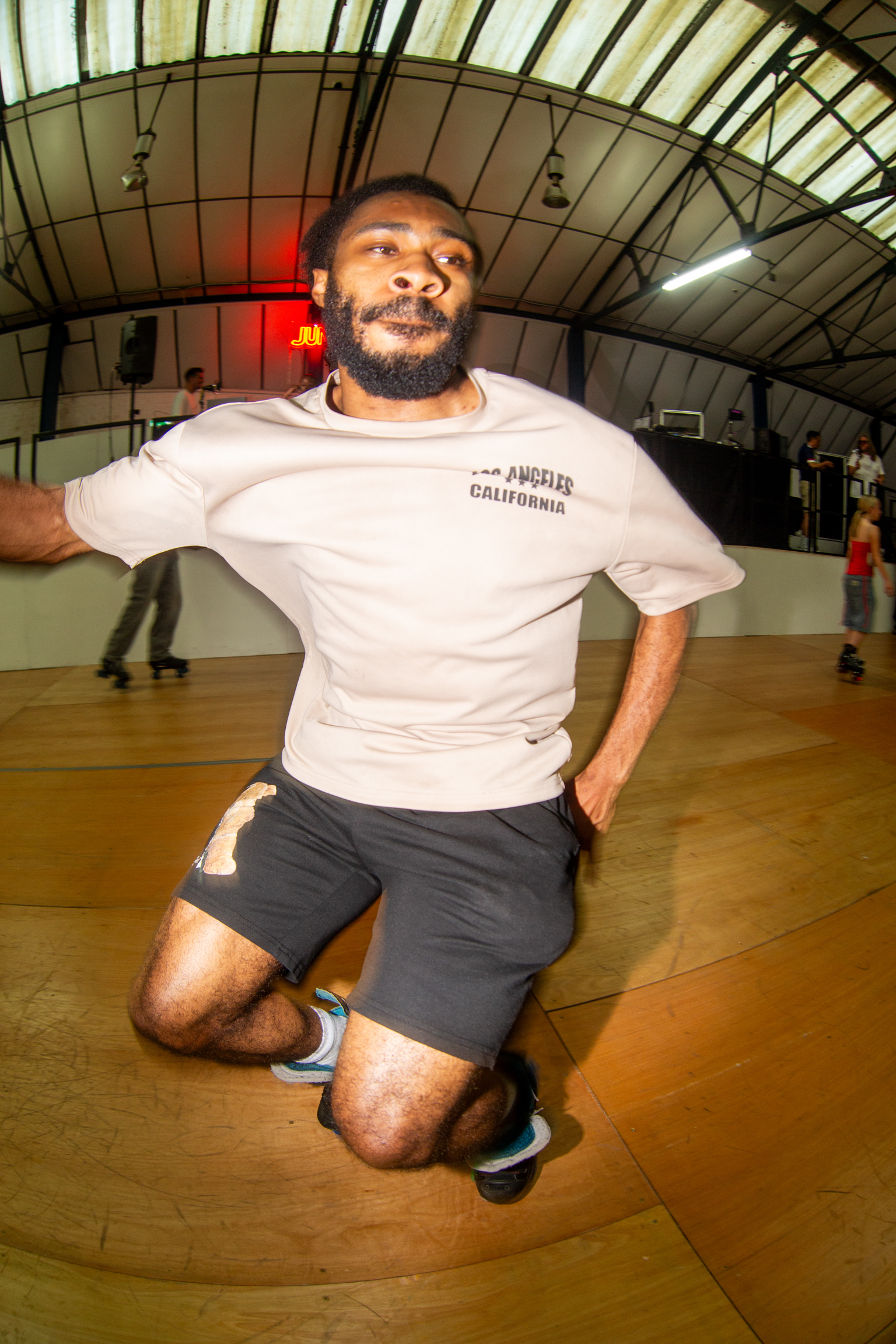 Man with beard kneeling on wooden floor in white Los Angeles California t-shirt and dark shorts inside curved glass-roofed building.