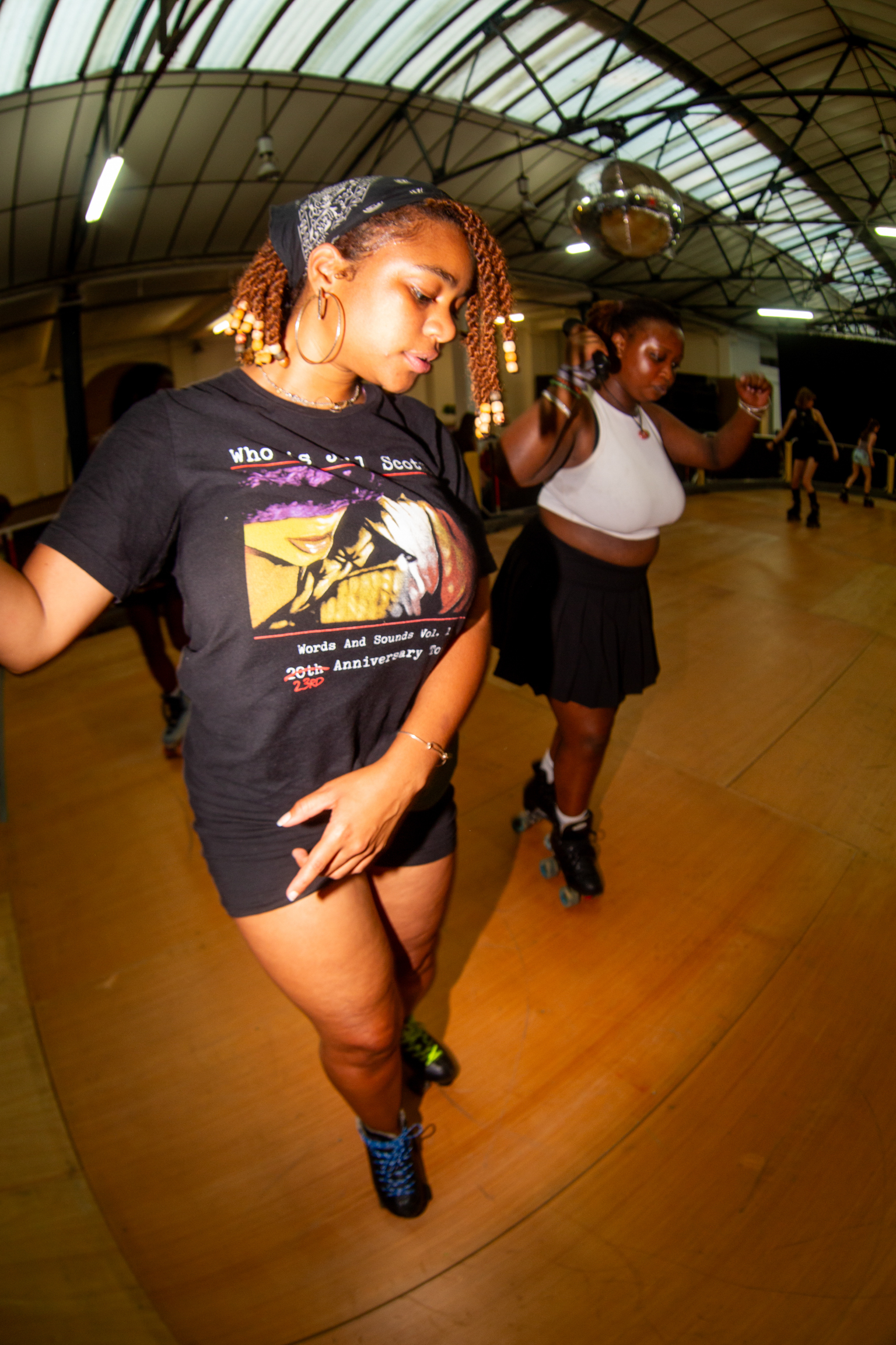 Woman with curly hair in black graphic t-shirt roller skating at indoor rink with arched ceiling and wooden floor.