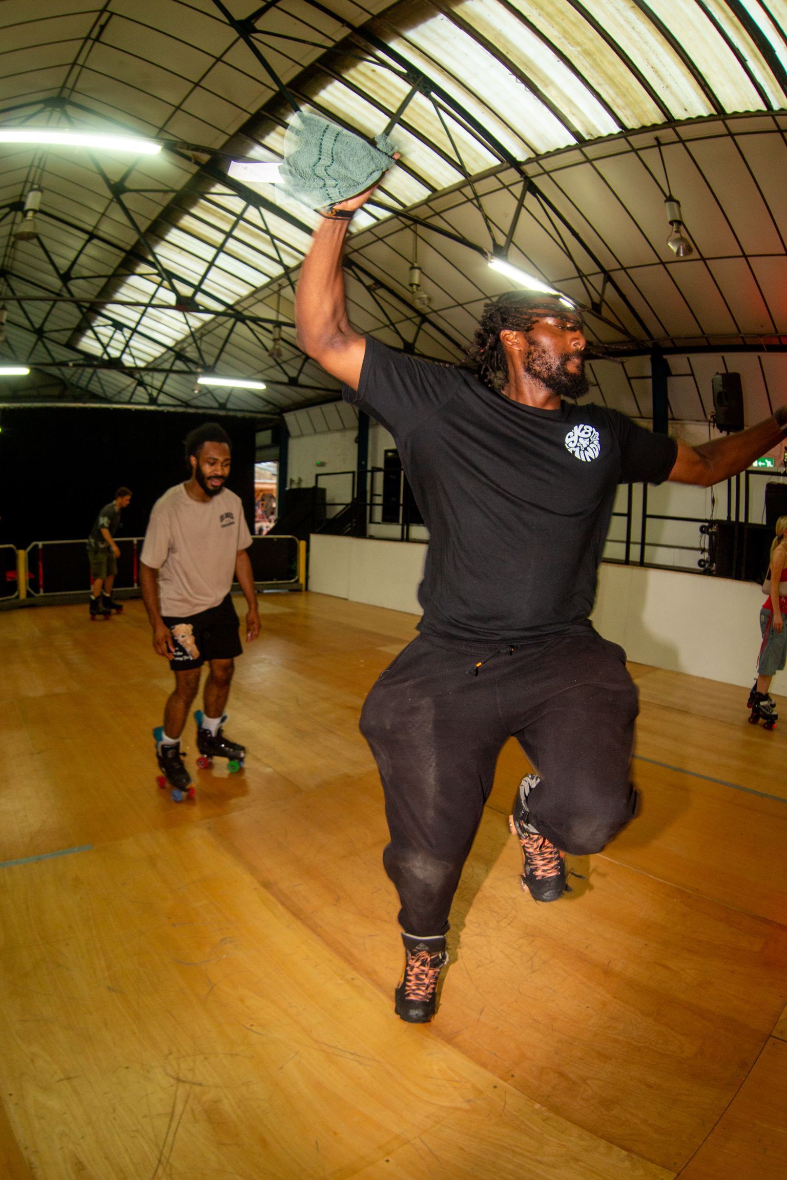Man roller skating indoors with arms raised, wearing black shirt and shorts. Others skate in background on wooden floor under arched ceiling.