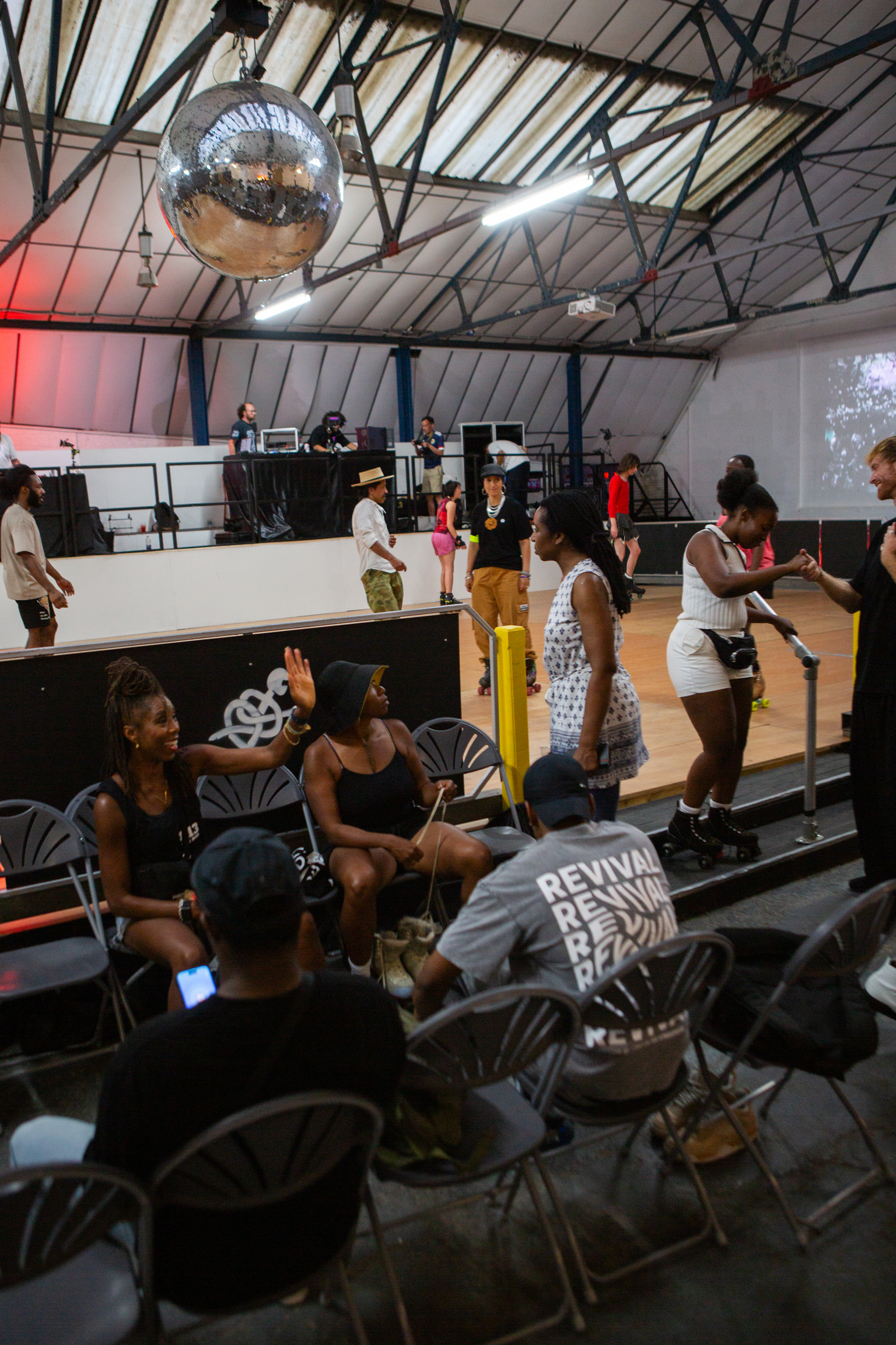 Indoor venue with exposed steel roof beams, disco ball, white raised platform with people dancing, audience seated on folding chairs below.