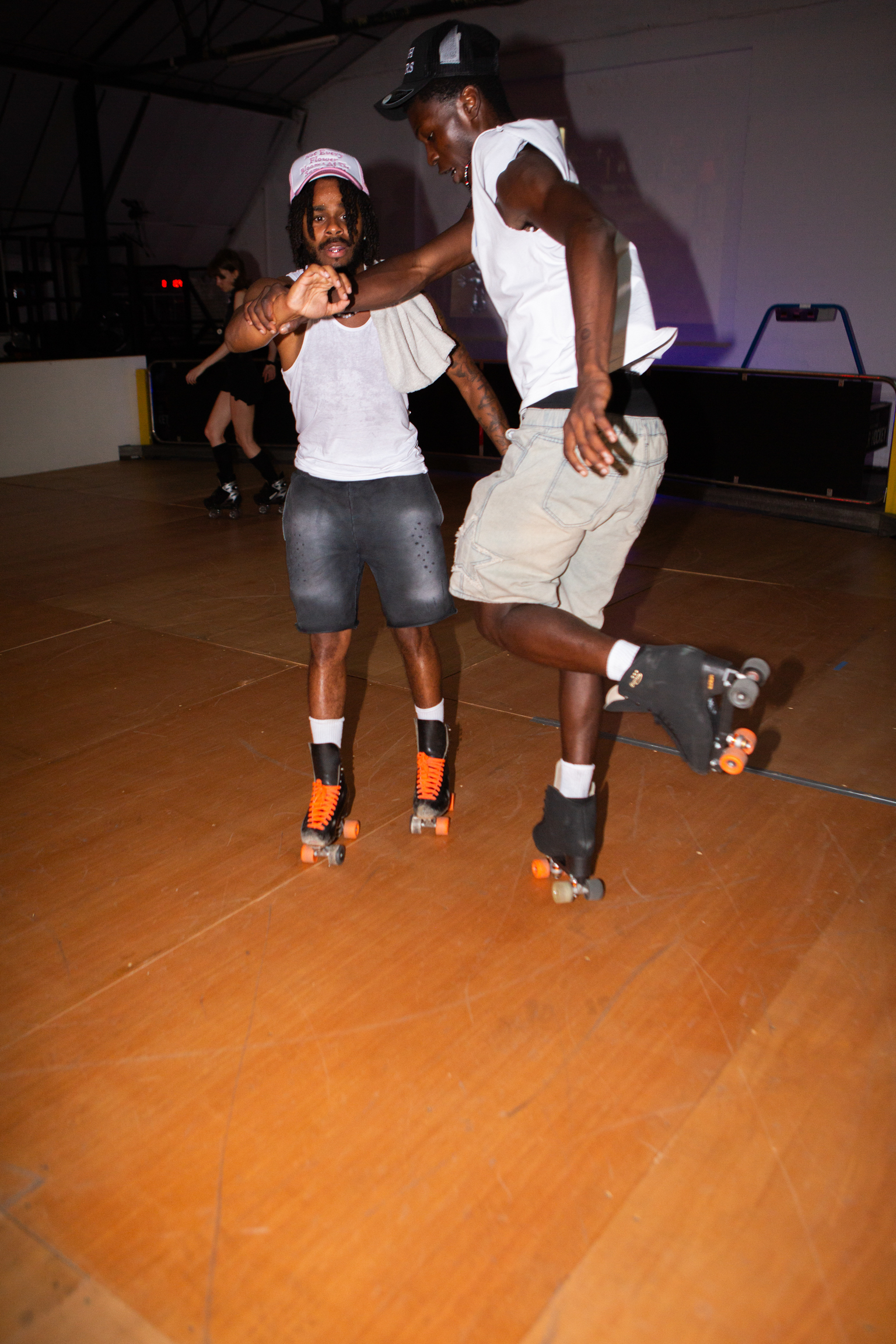 Two people roller skating together on wooden floor, wearing white tops and caps with orange and black roller skates.