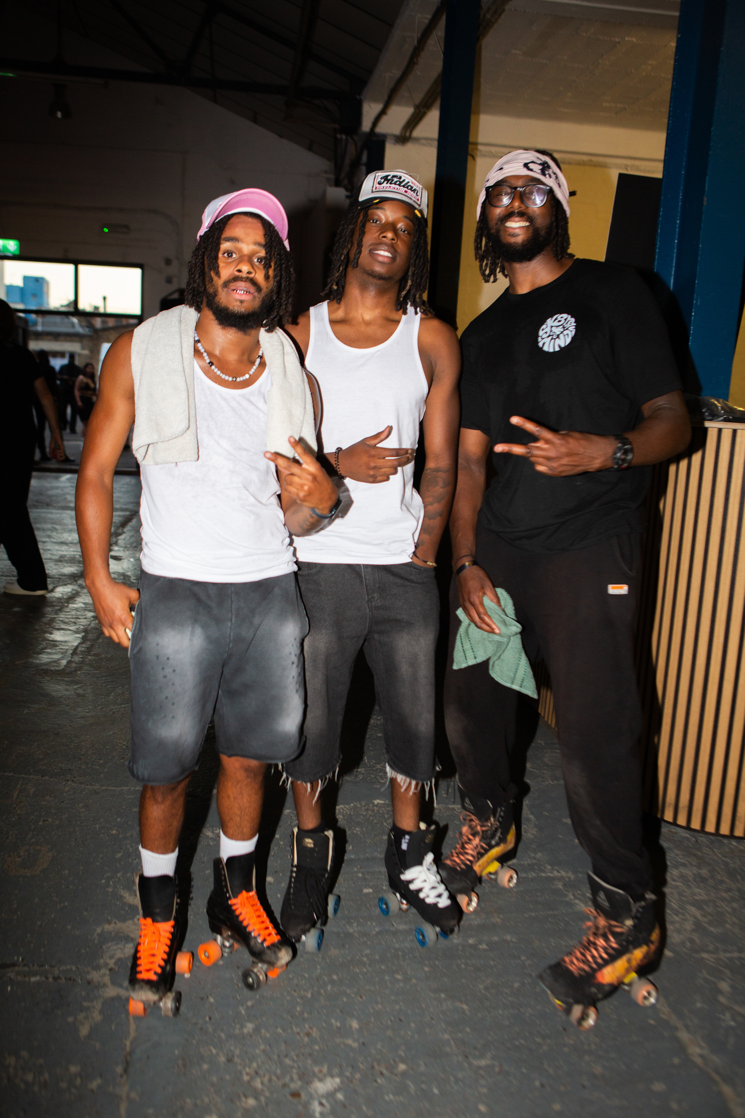 Three men posing together at roller skating rink wearing white vests, caps, and colourful roller skates on dark floor.