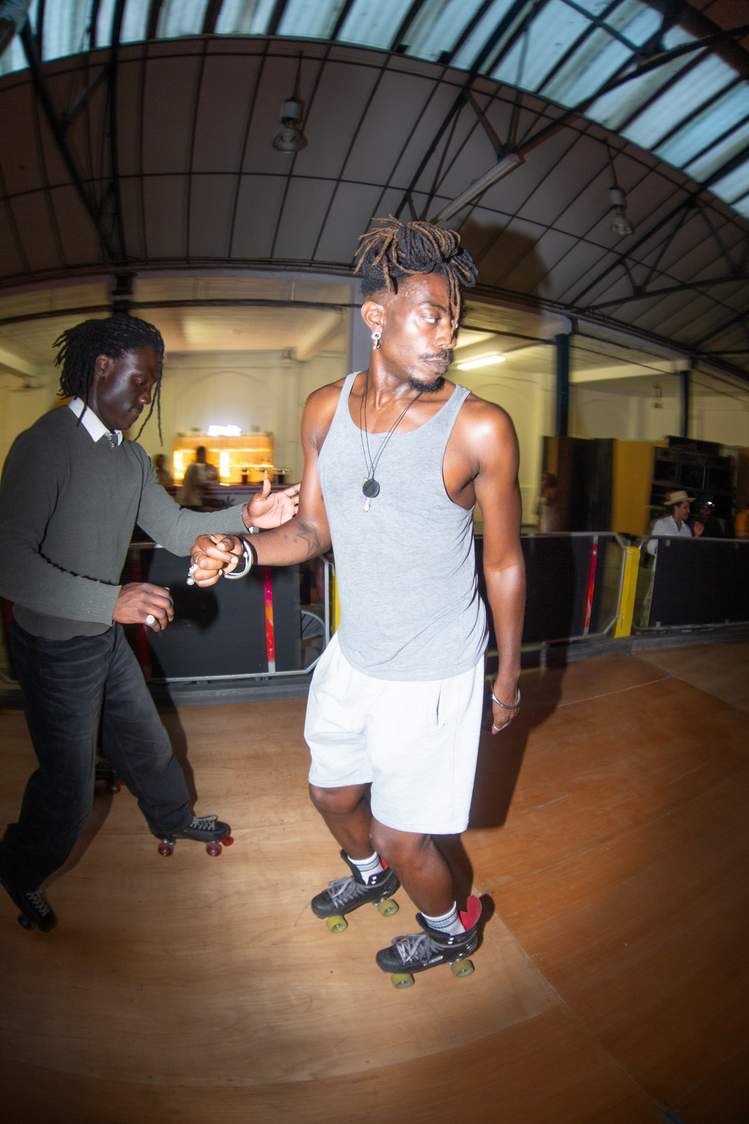Two people roller skating in indoor rink with arched glass ceiling, one wearing grey vest and white shorts, other in dark clothing.