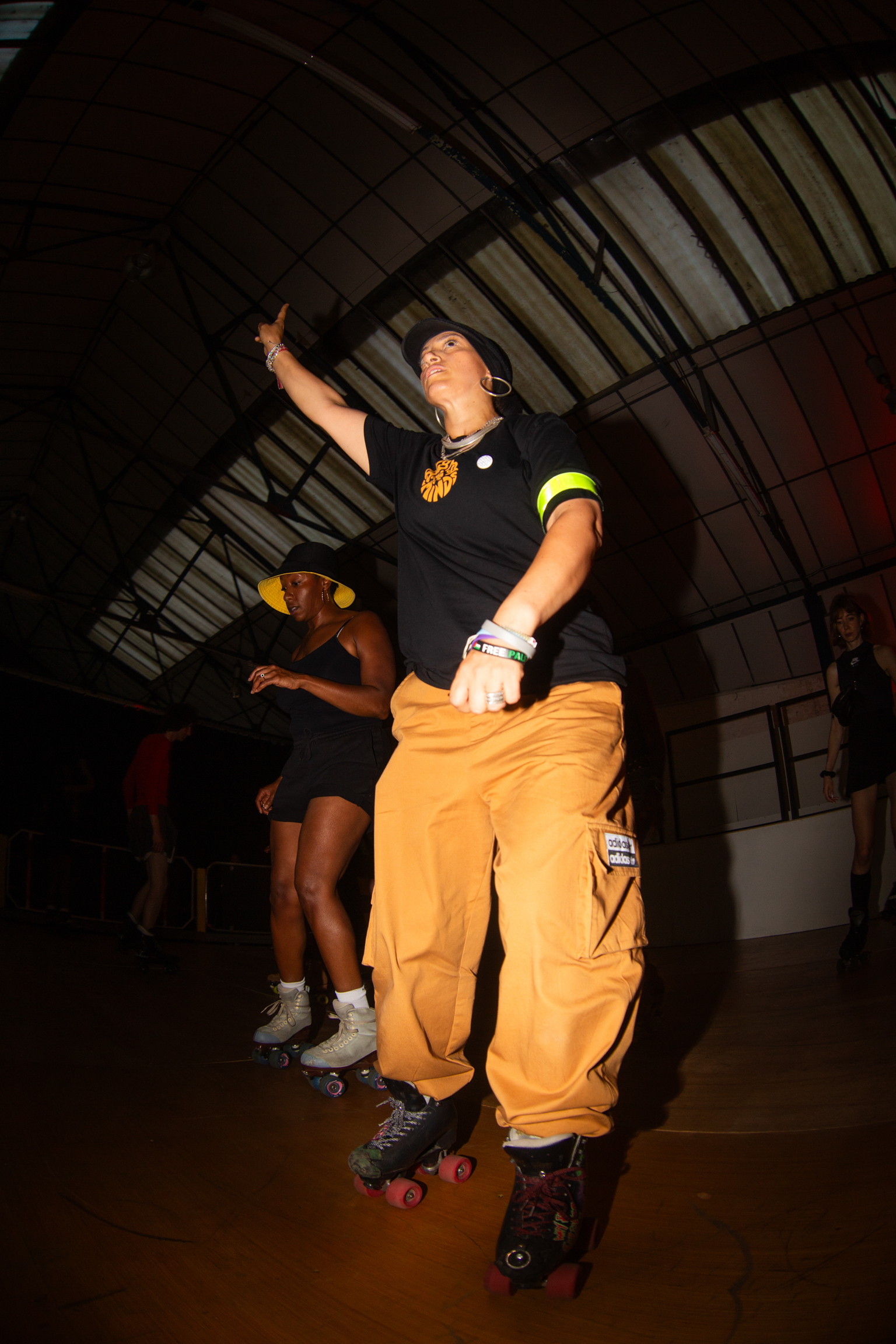 Person in black top and orange baggy trousers roller skating indoors, arm raised, with others skating in background under metal roof.