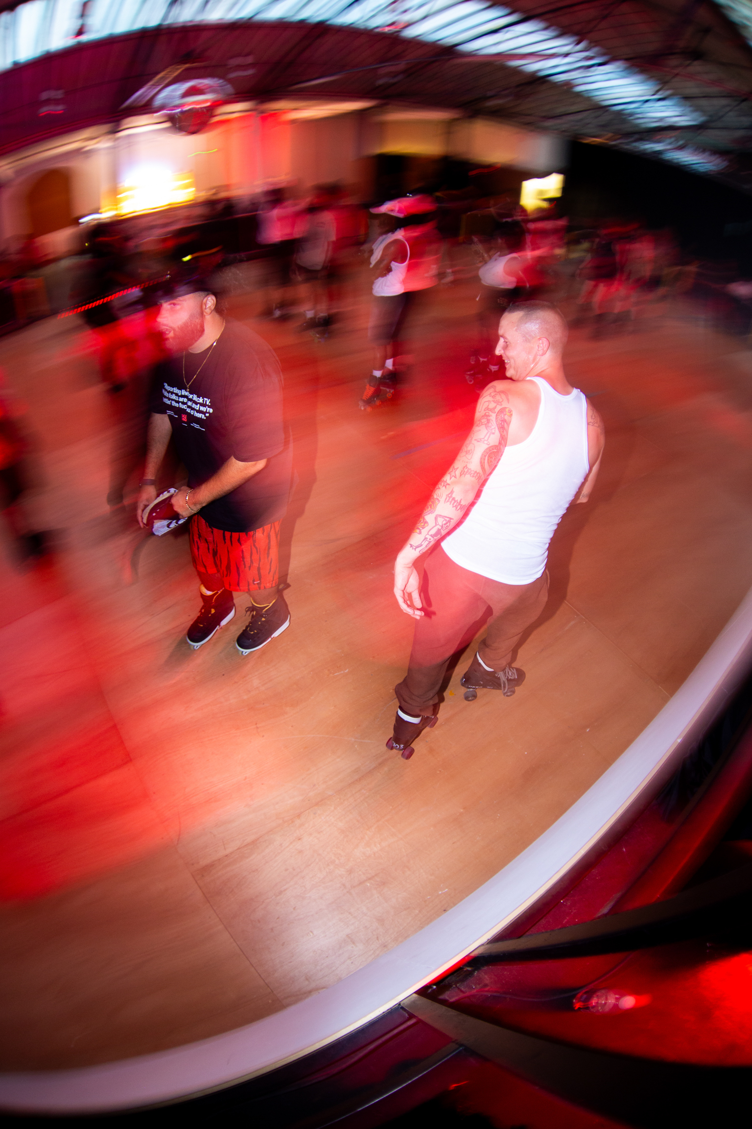 Blurred motion image of people dancing on wooden floor with red lighting and curved architectural elements overhead.