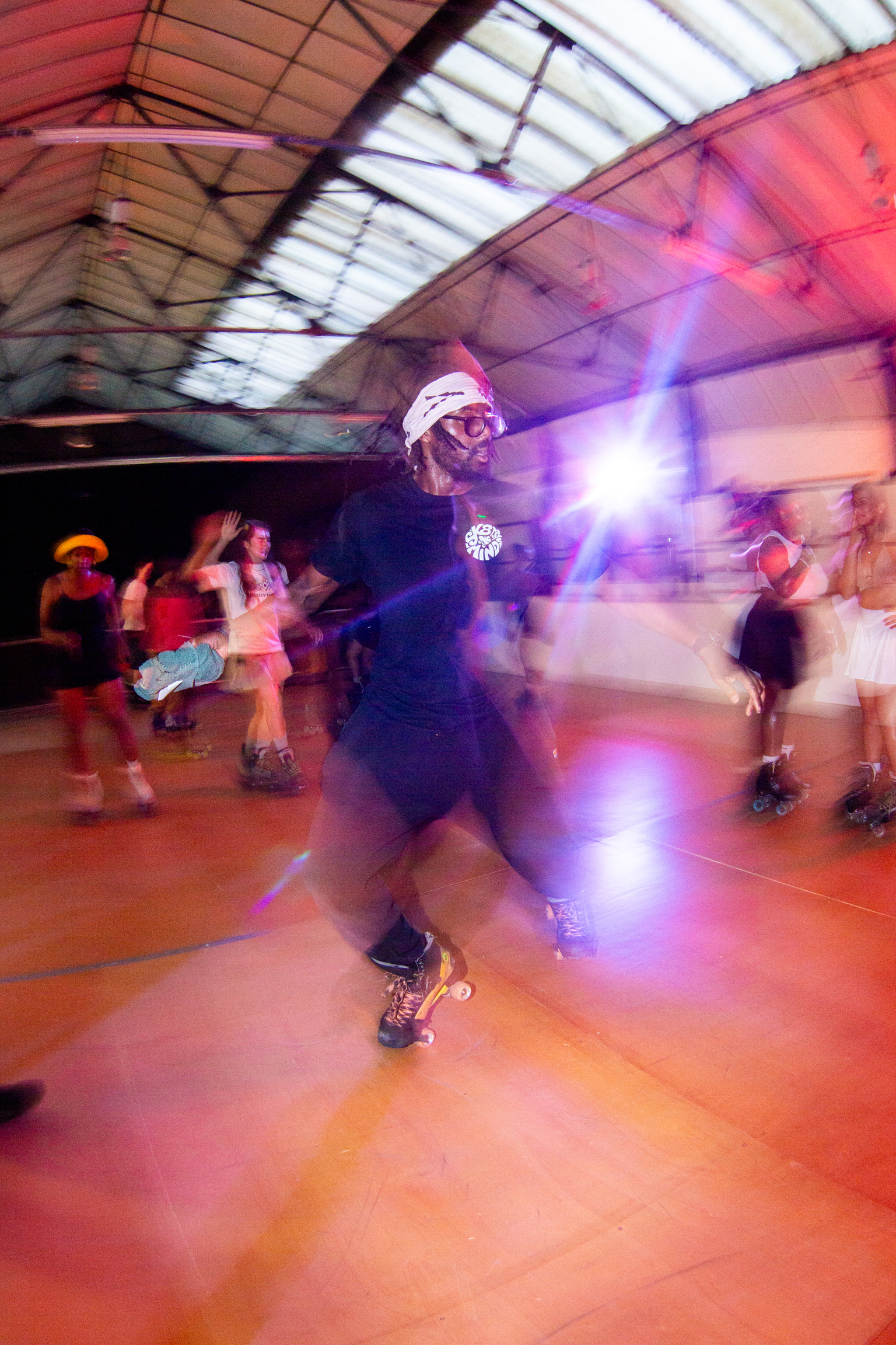 Motion-blurred roller skaters in indoor rink with red lighting and glass roof. Central figure in white hat, multiple people skating in background.