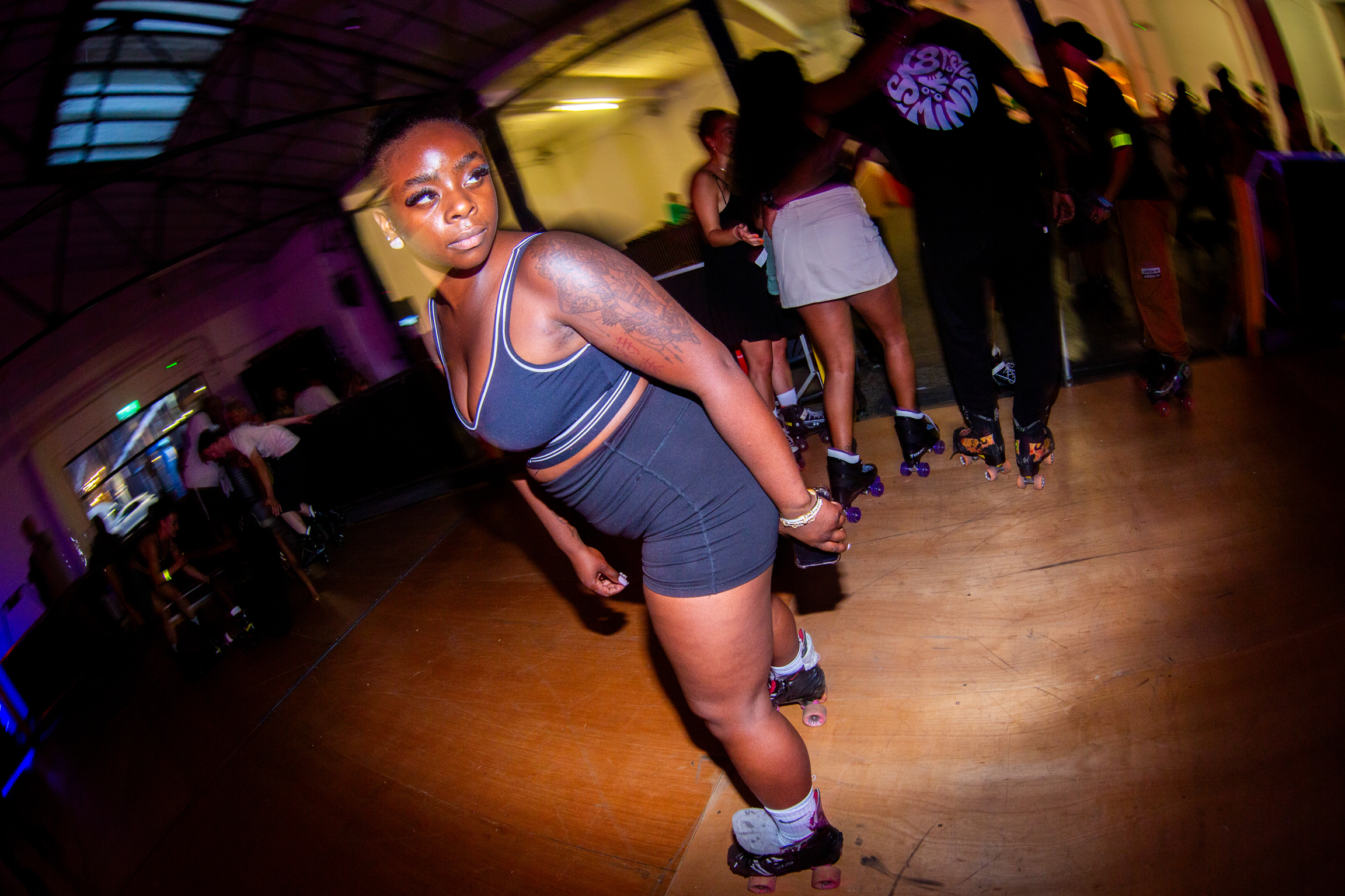 Woman in grey athletic wear roller skating on wooden floor in dimly lit venue with other skaters in background.