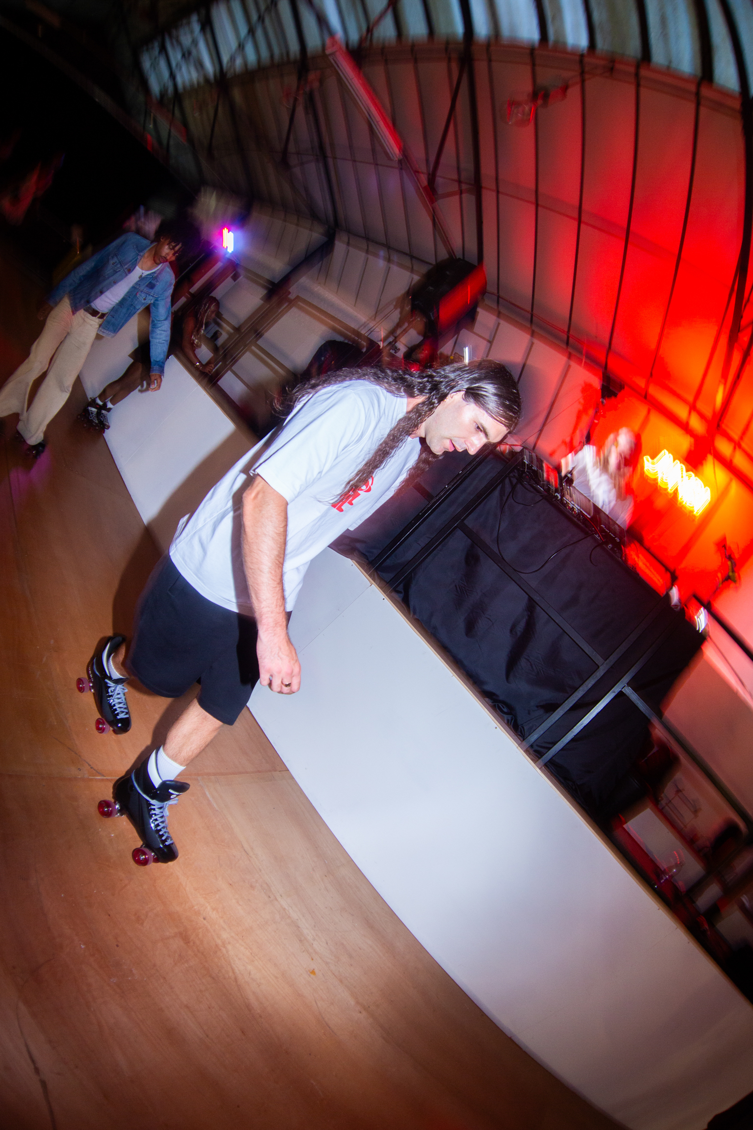 Person on roller skates leaning forward on white ramp in dimly lit skate park with red lighting and metal barriers.