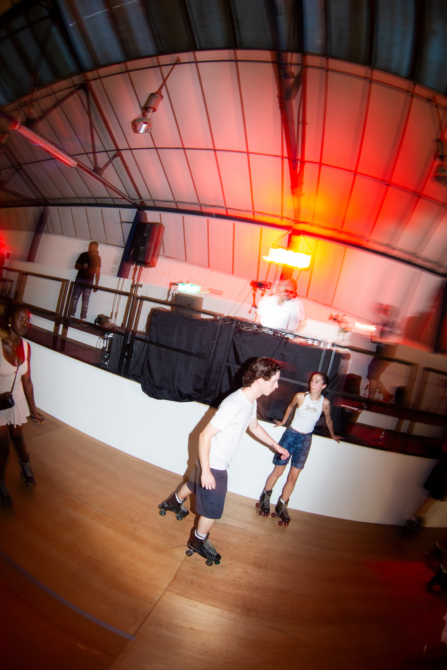 Two people roller skating on white rink with wooden floors, red lighting overhead, curved ceiling with metal framework visible above.