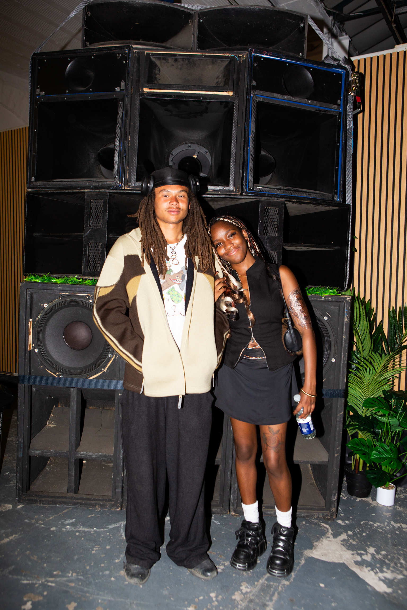 Two people posing in front of large black speaker stack. Person on left wears beanie and beige hoodie, person on right wears black outfit.
